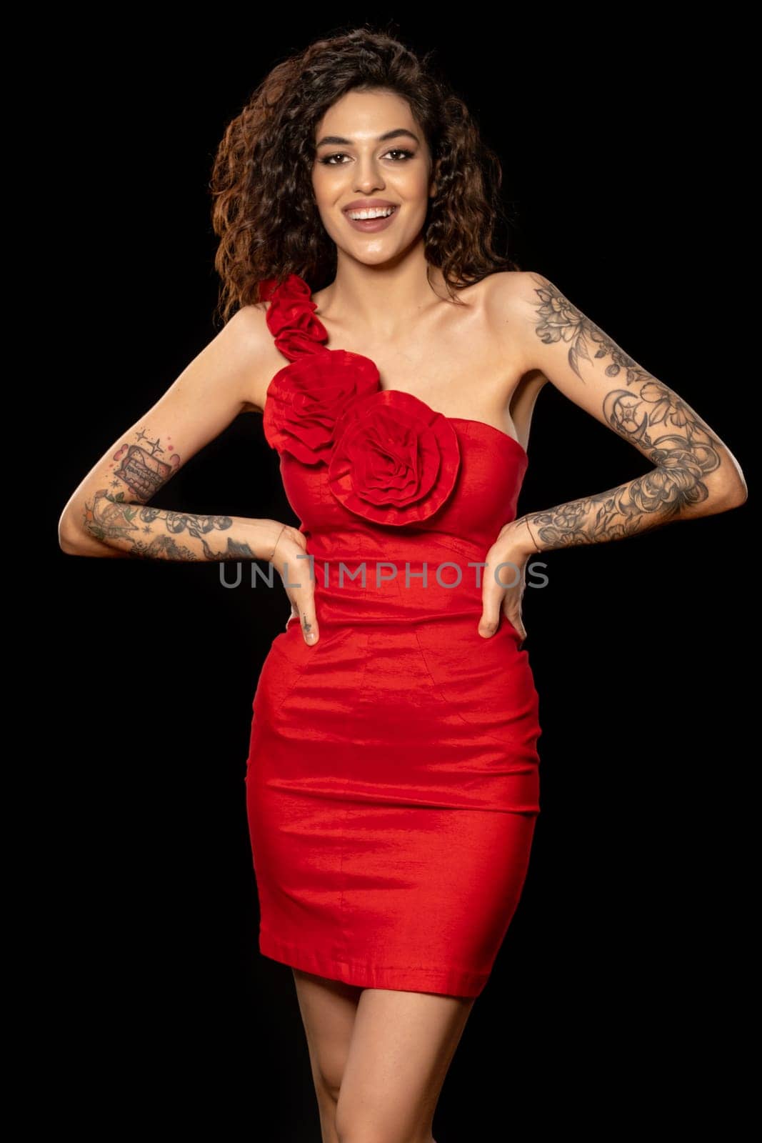 Cheerful excited young brunette with tattooed arms, wearing red cocktail dress posing on dark studio background, standing akimbo and laughing, looking at camera