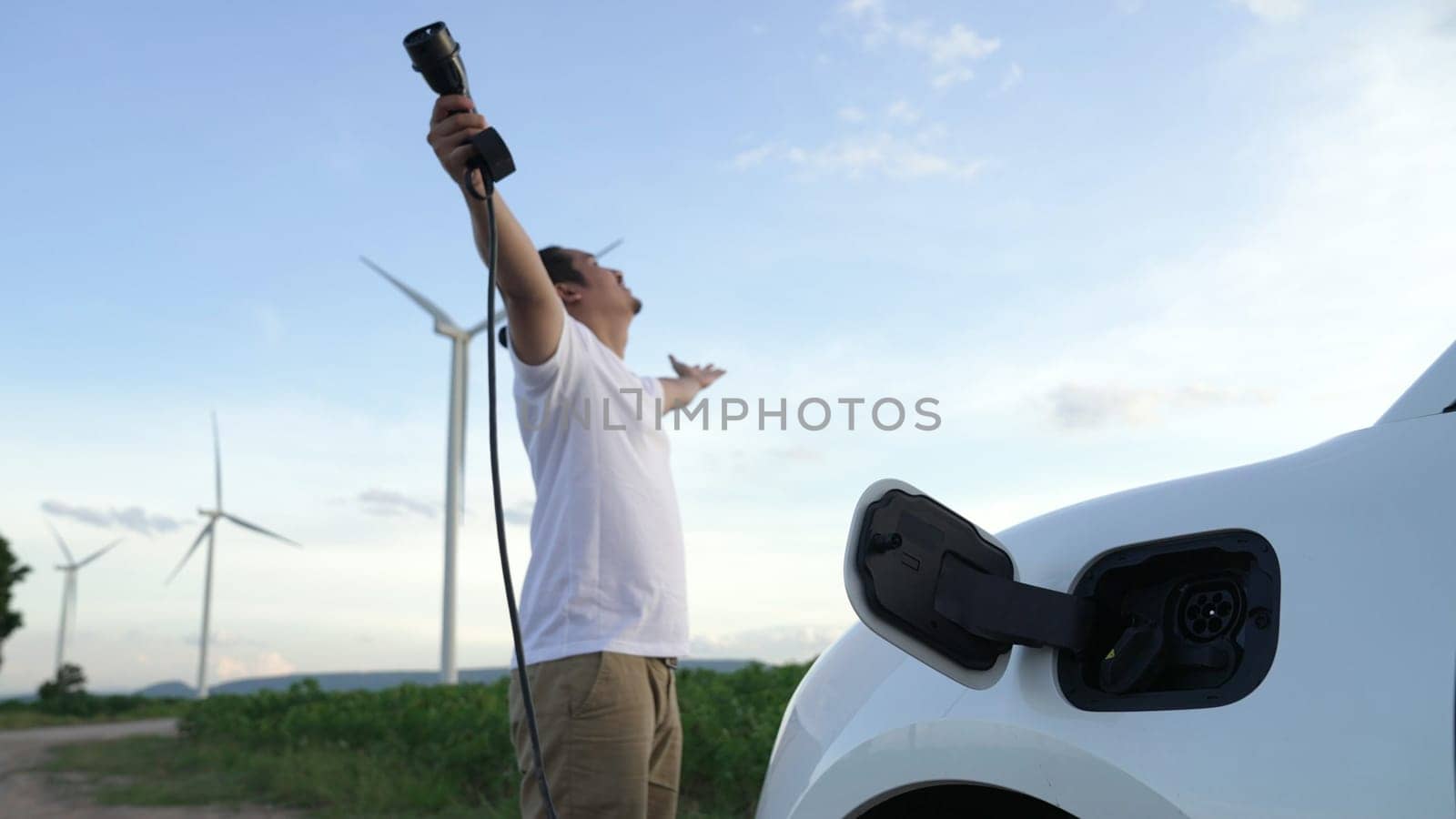 Progressive man with his electric car, EV car recharging energy from charging station on green field with wind turbine as concept of future sustainable energy. Electric vehicle with energy generator.