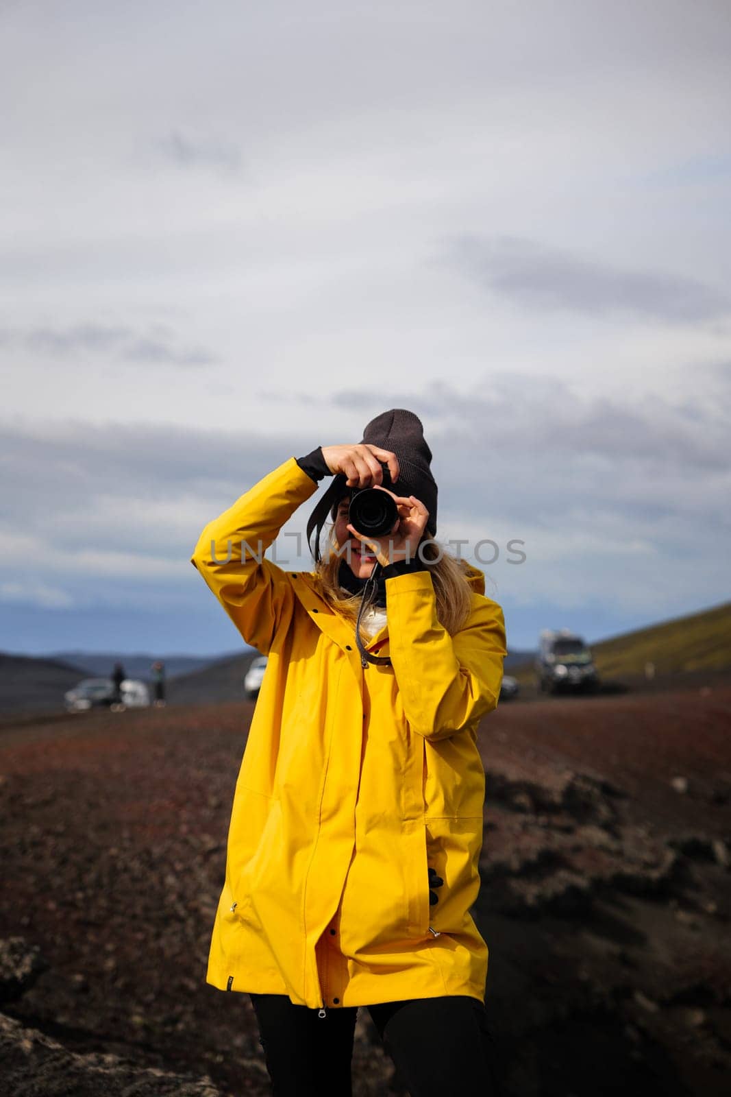 A backpacking girl taking a photo of her surroundings in the Icelandic highlands. She is wearing a yellow trekking jacket and faces the camera while holding her camera at eye level. Travel, outdoor, and nature photography. Blahylur lake, Iceland.