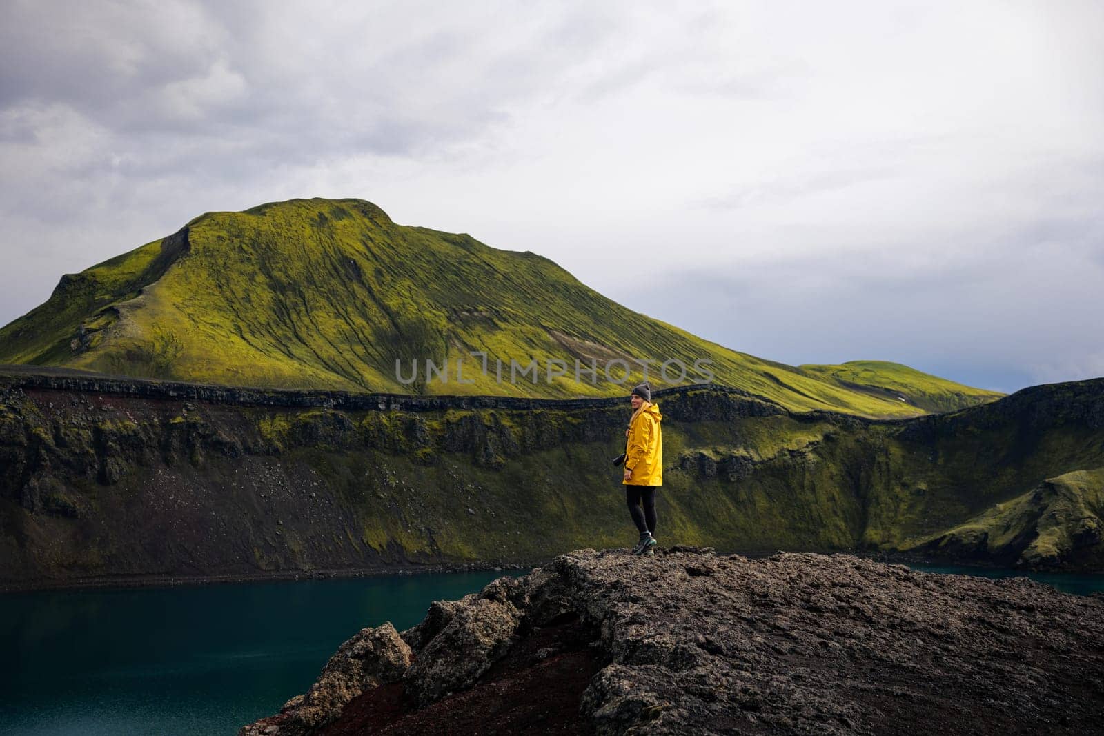 This photo shows a backpacker girl smiling at the view. She is wearing a yellow trekking jacket and a hat, and she has a camera slung over her shoulder. The sky is cloudy, and the background is a mountain range. This photo is perfect for use in travel and nature photography. It could be used to illustrate a story about hiking in the mountains, or it could simply be used to create a sense of happiness and contentment.