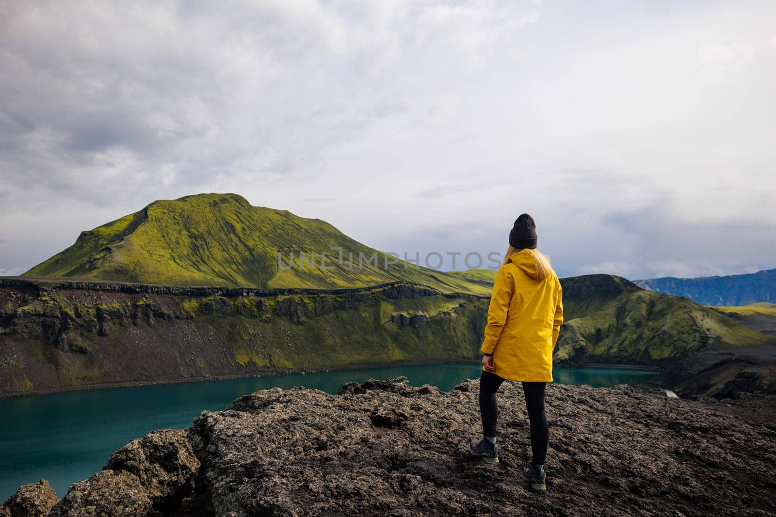 This photo shows a backpacker girl enjoying the beauty of Iceland. She is standing on the shore of Blahylur lake, looking out at the surrounding mossy mountains. The sky is cloudy. Travel and nature photography. It could be used to illustrate a story about the beauty of Iceland, or it could simply be used to create a sense of wonder. Blahylur lake, Iceland highlands.