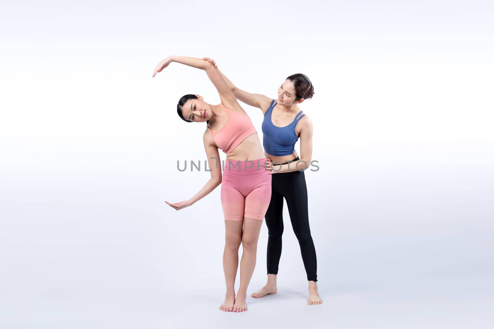 Asian woman in sportswear doing yoga exercise in standing pose with her trainer or yoga buddy. Healthy body care and meditation yoga lifestyle in full shot on isolated background. Vigorous