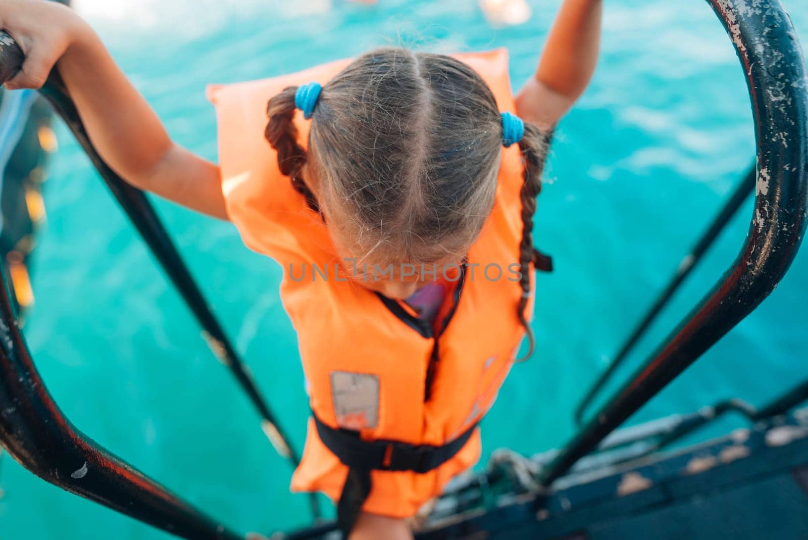 A young girl, sporting a bright orange life jacket, climbs a boat ladder over the vibrant turquoise waters