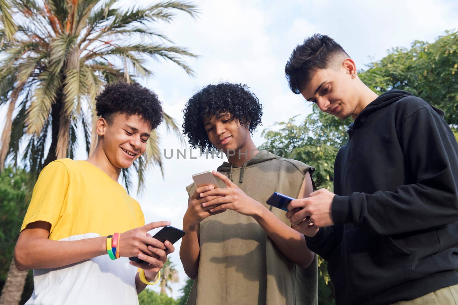 low angle of teen diverse friends having fun surfing Internet on cellphones smiling happy in park, concept of youth lifestyle and technology of communication
