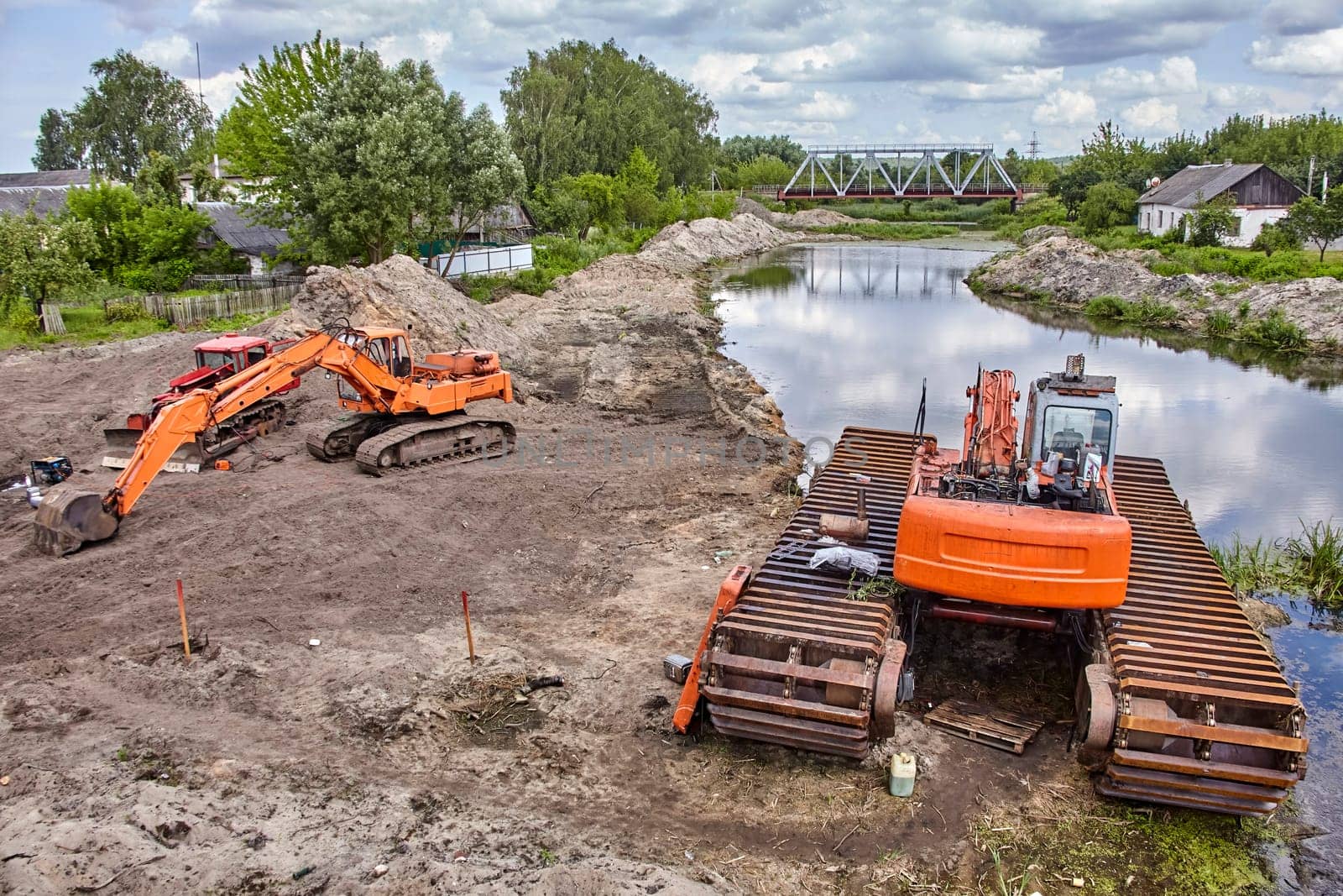 A bright orange excavator sits beside a calm river, its tracks resting on the sandy bank. The river winds its way under a metal bridge in the distance, passing through a lush green landscape.