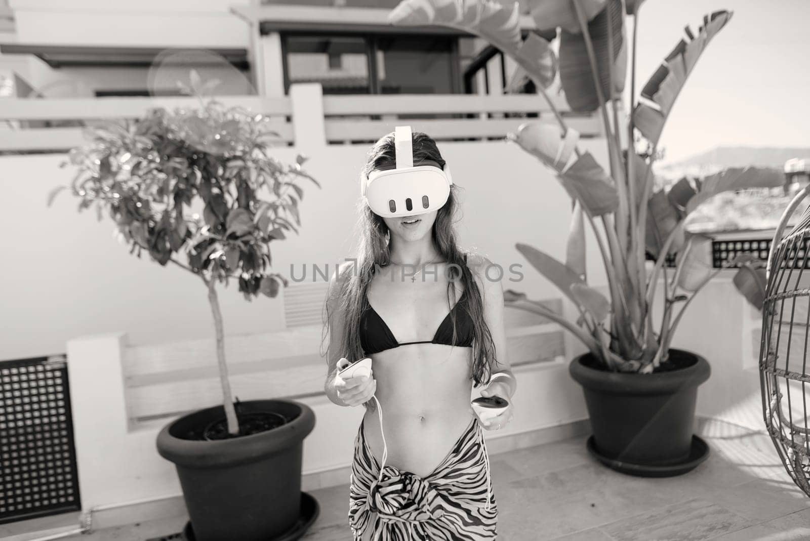 A young woman in a stylish bikini is enjoying a VR headset in a chic outdoor area with vibrant, lush plants