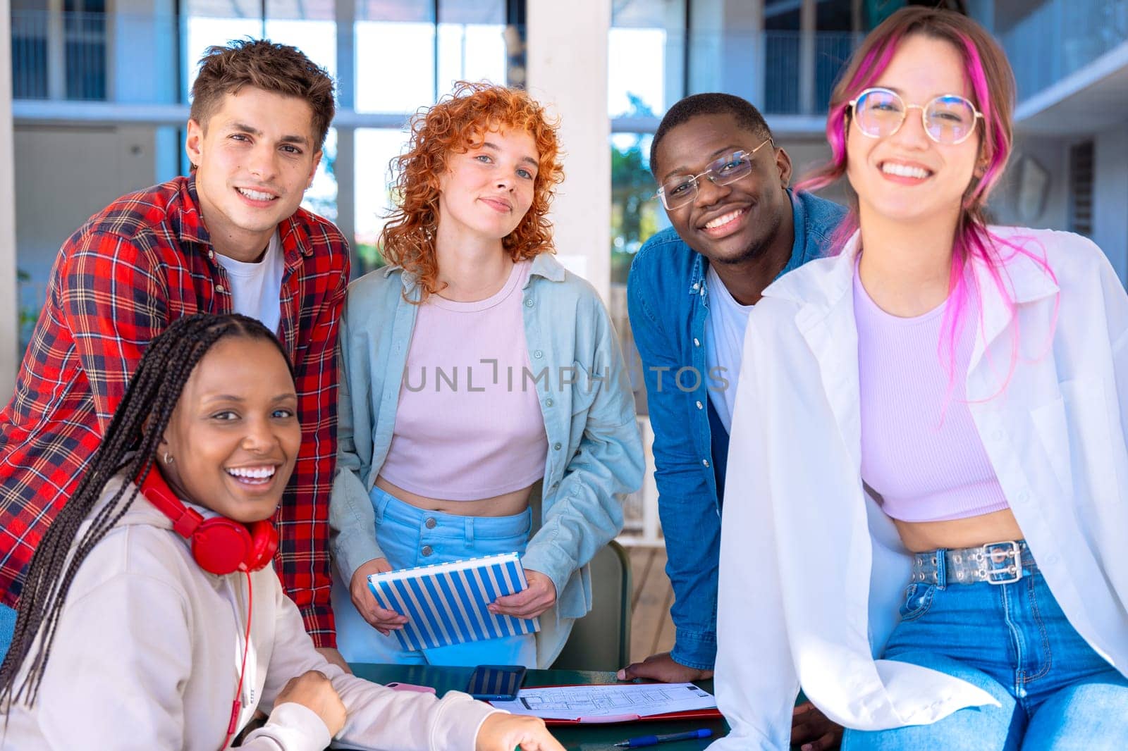 A group of five diverse students studying and collaborating at a library table working on a shared project, with books and devices on the table.