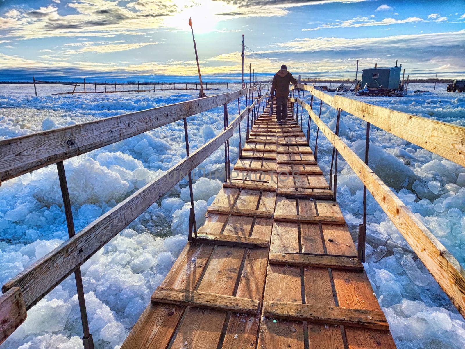 A wooden bridge stretches over frozen waters with snow and structures in the background during early morning light.