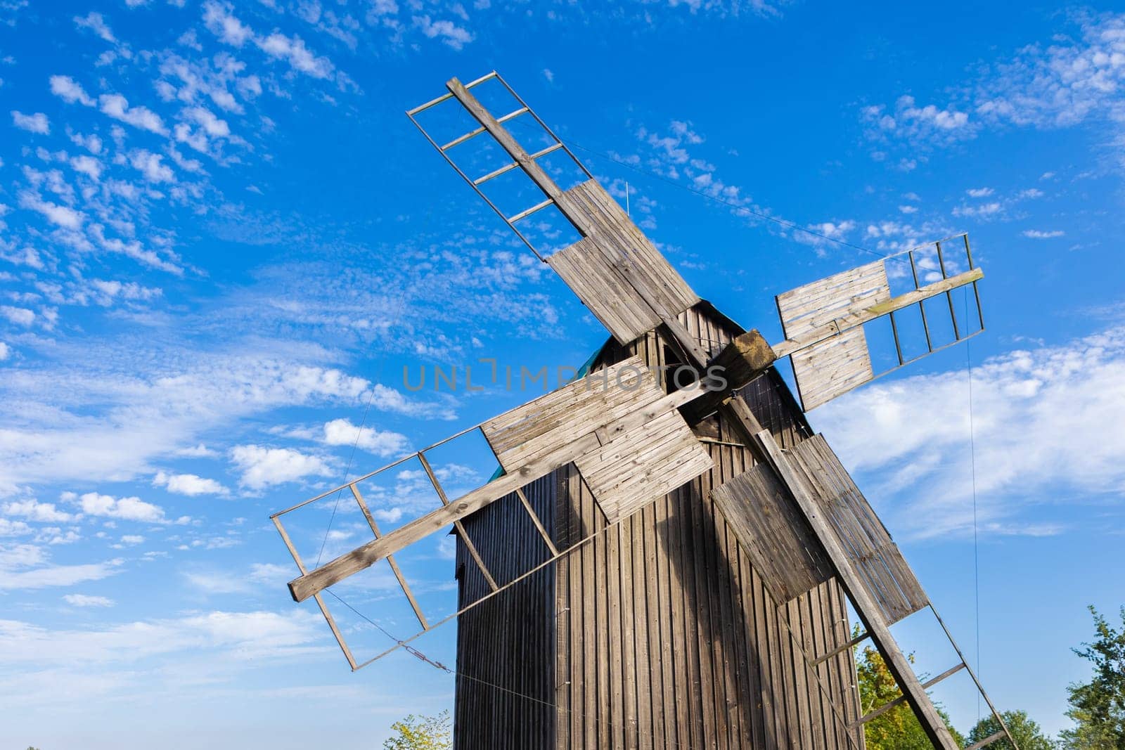 Traditional wooden windmill under a bright blue sky, with scattered clouds. The weathered wood and simple construction of the windmill evoke a rustic, historical feel, representing rural life and sustainable energy from a bygone era.