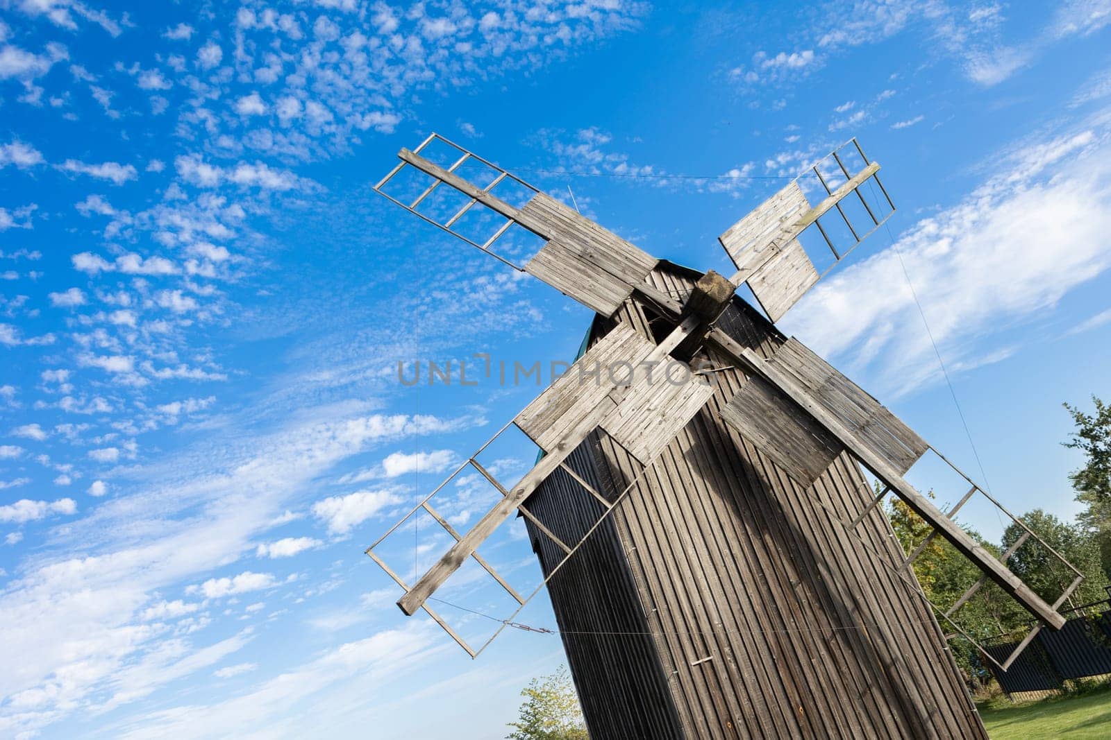 Traditional wooden windmill under a bright blue sky, with scattered clouds. The weathered wood and simple construction of the windmill evoke a rustic, historical feel, representing rural life and sustainable energy from a bygone era.