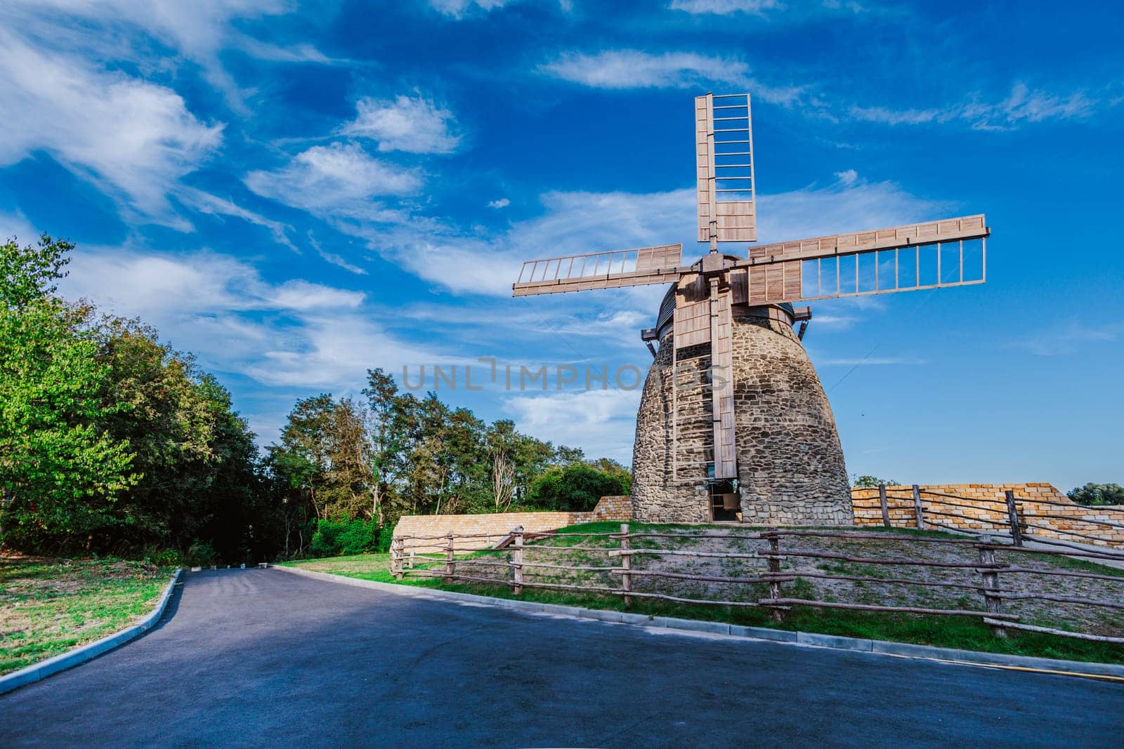 Stone windmill stands tall under a vibrant blue sky with scattered clouds. The wooden blades of the windmill stretch outwards, capturing the essence of traditional rural life and old-world architecture.