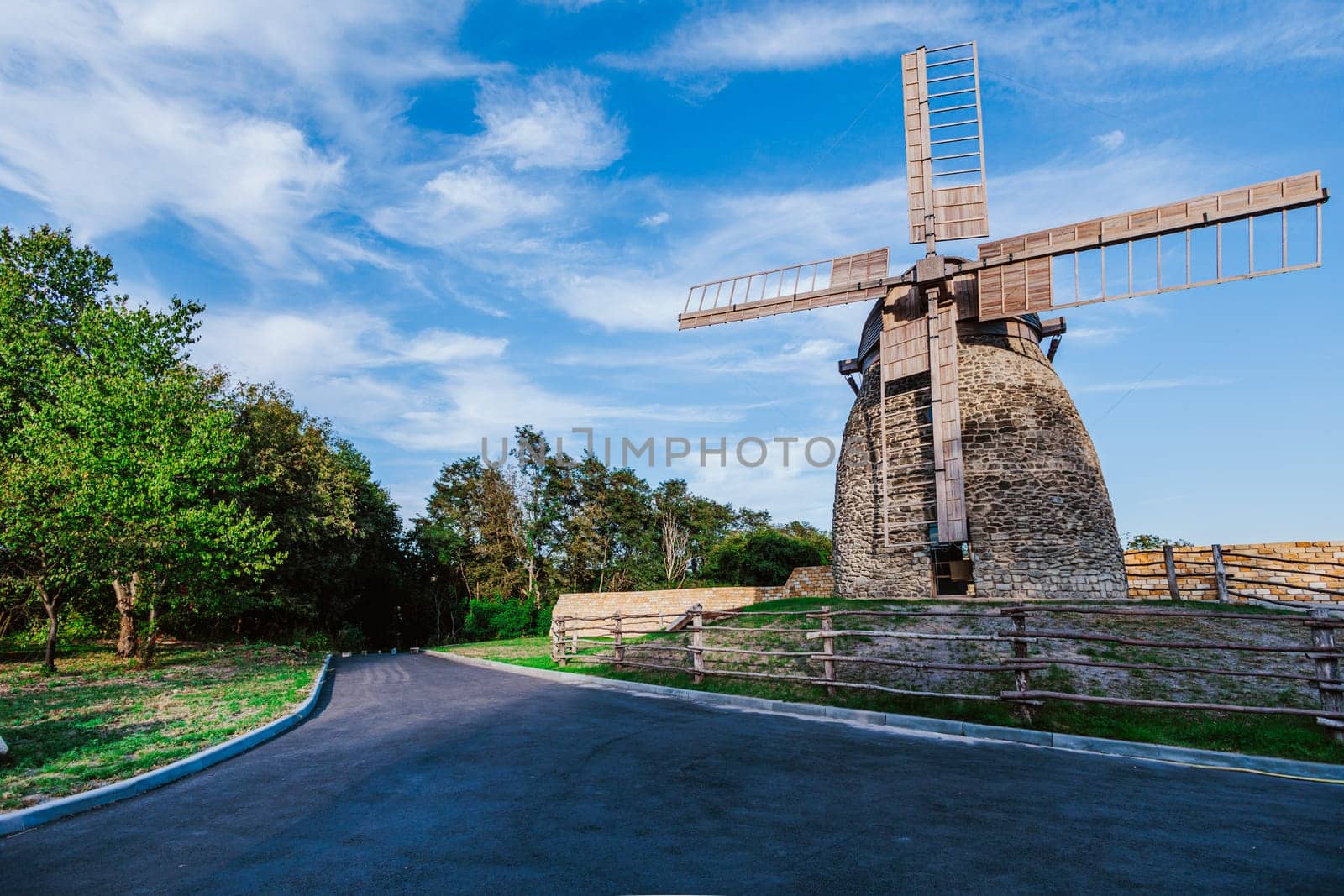 Stone windmill stands tall under a vibrant blue sky with scattered clouds. The wooden blades of the windmill stretch outwards, capturing the essence of traditional rural life and old-world architecture.