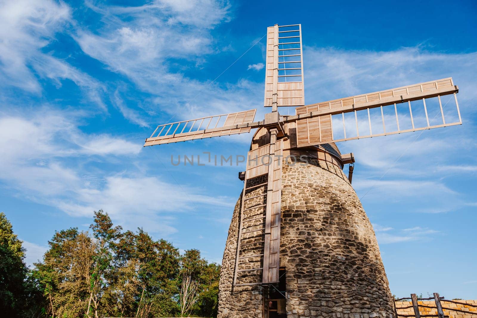 Old stone windmill stands against a clear blue sky with gentle clouds. The windmill wooden blades and stone structure showcase traditional craftsmanship and the historical significance of rural architecture.