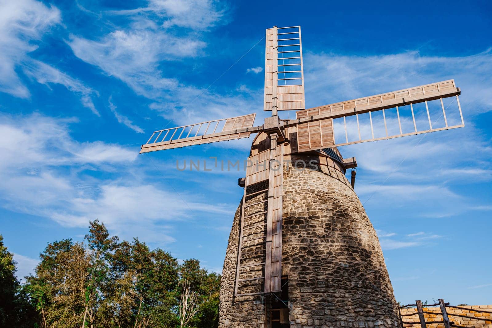 Old stone windmill stands against a clear blue sky with gentle clouds. The windmill wooden blades and stone structure showcase traditional craftsmanship and the historical significance of rural architecture.