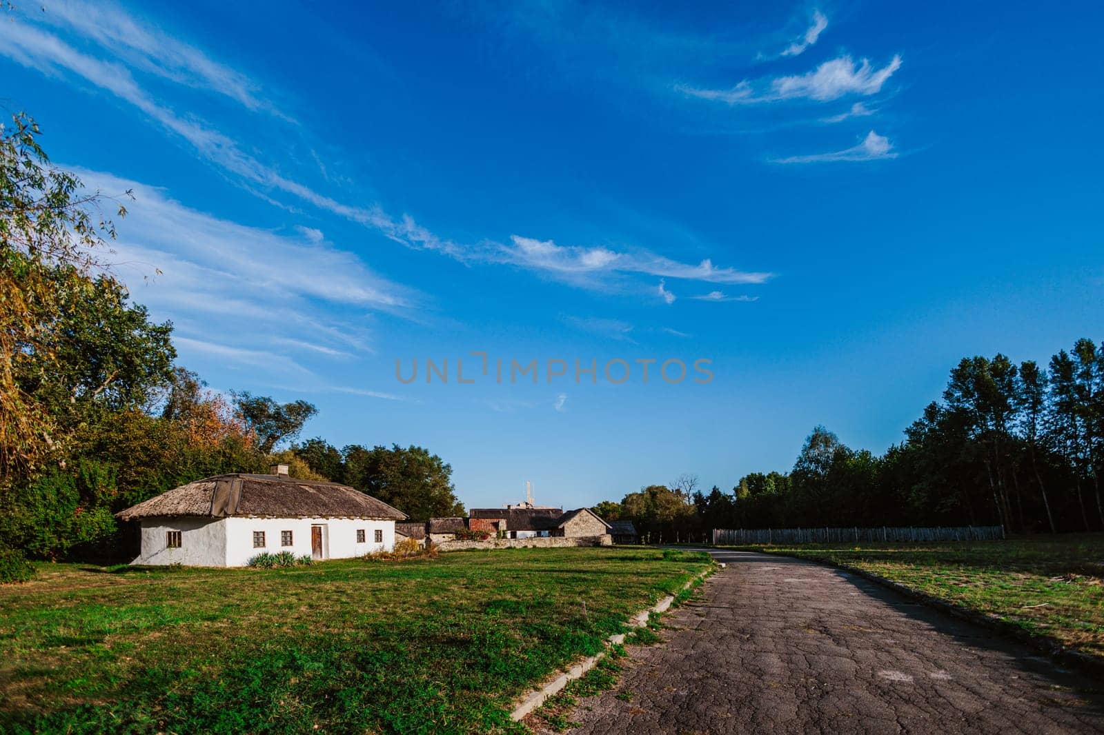 Peaceful rural scene featuring a traditional thatched-roof house and a wide open road under a vast blue sky. The whitewashed house stands out against the greenery of trees and fields, creating a picturesque countryside setting.
