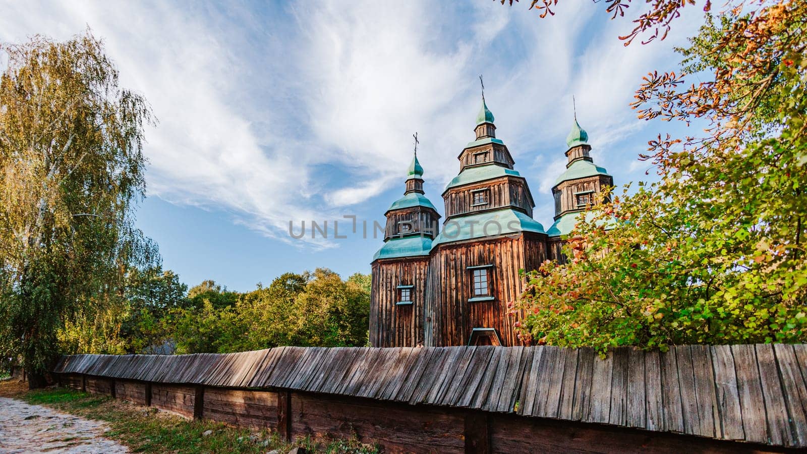 Traditional wooden church with green domes rises behind a rustic wooden fence, set against a beautiful blue sky with wispy clouds.