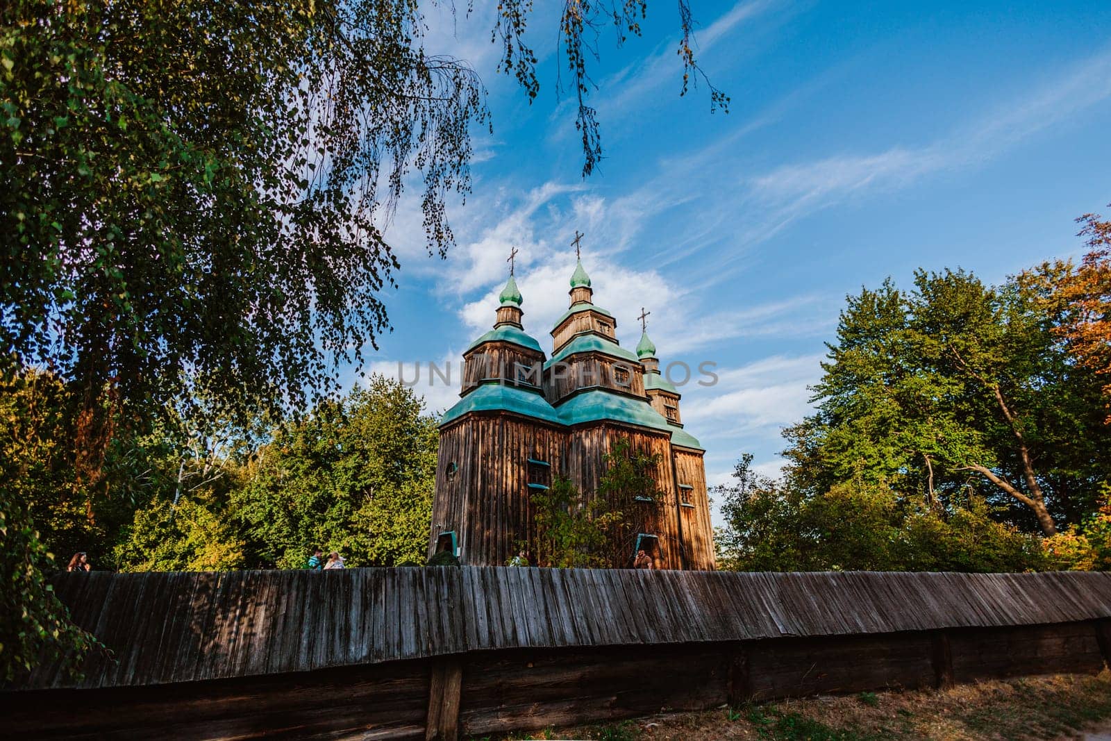 Traditional wooden church with green domes rises behind a rustic wooden fence, set against a beautiful blue sky with wispy clouds.