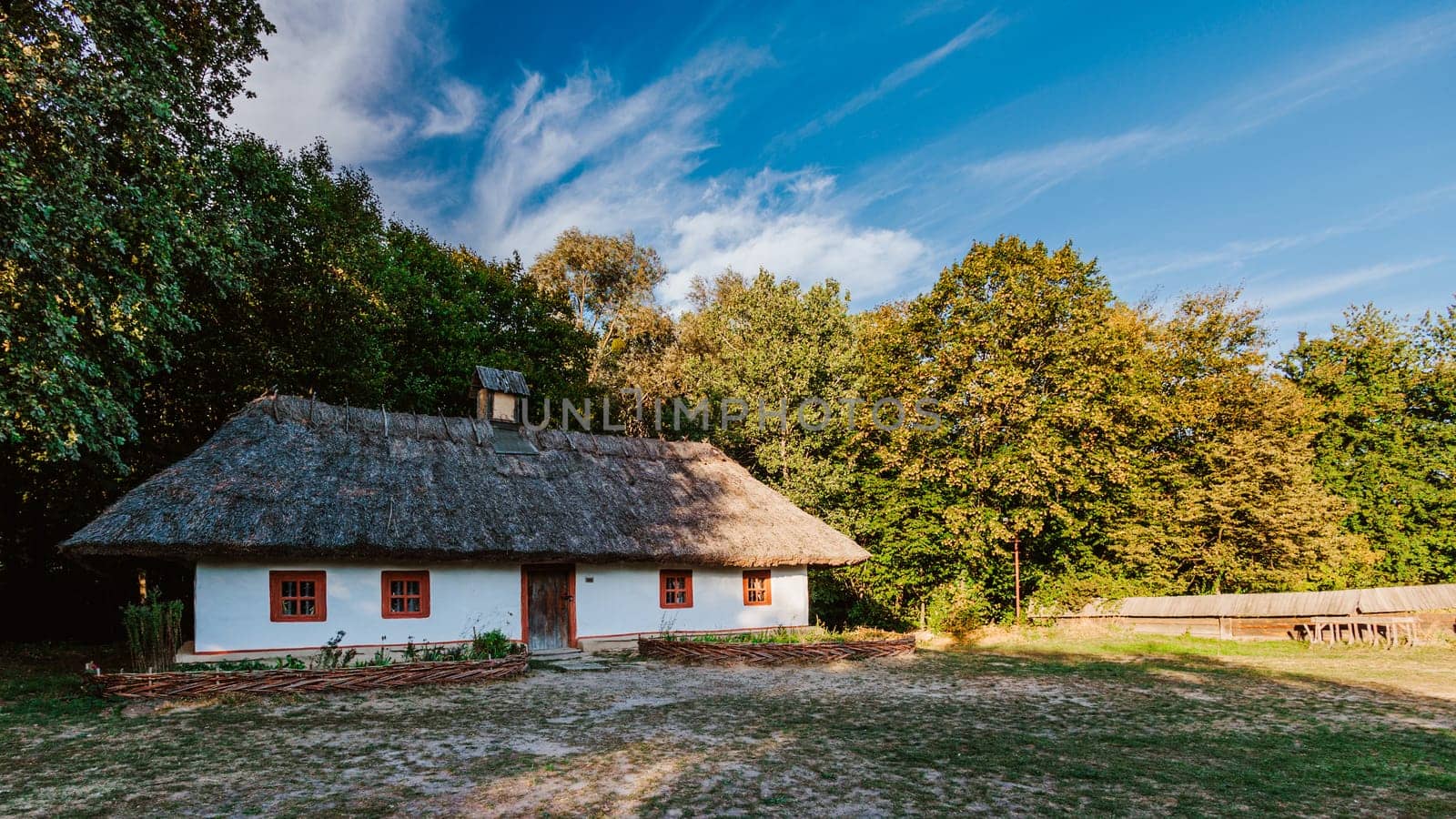 Traditional thatched-roof house sits peacefully in a rural setting, surrounded by lush greenery under a clear blue sky. The whitewashed walls and rustic wooden windows emphasize the simplicity and beauty of countryside life.