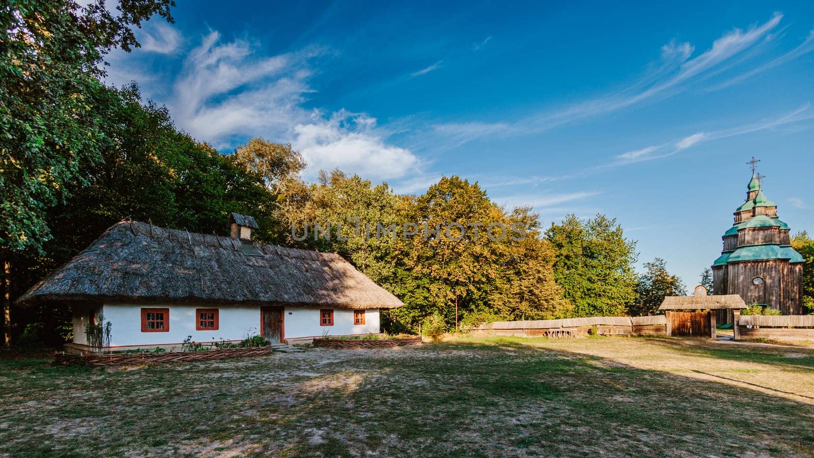 Scene featuring a traditional thatched-roof house with whitewashed walls, surrounded by greenery under a clear blue sky. In the distance, a historical wooden church with green domes adds cultural and architectural depth to the scene.
