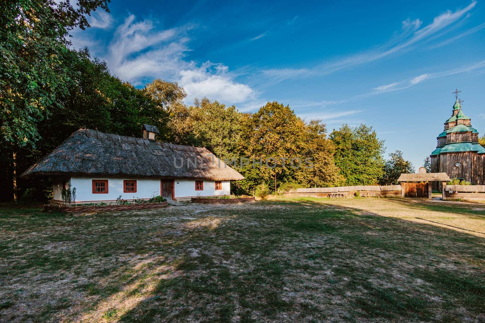 Scene featuring a traditional thatched-roof house with whitewashed walls, surrounded by greenery under a clear blue sky. In the distance, a historical wooden church with green domes adds cultural and architectural depth to the scene.