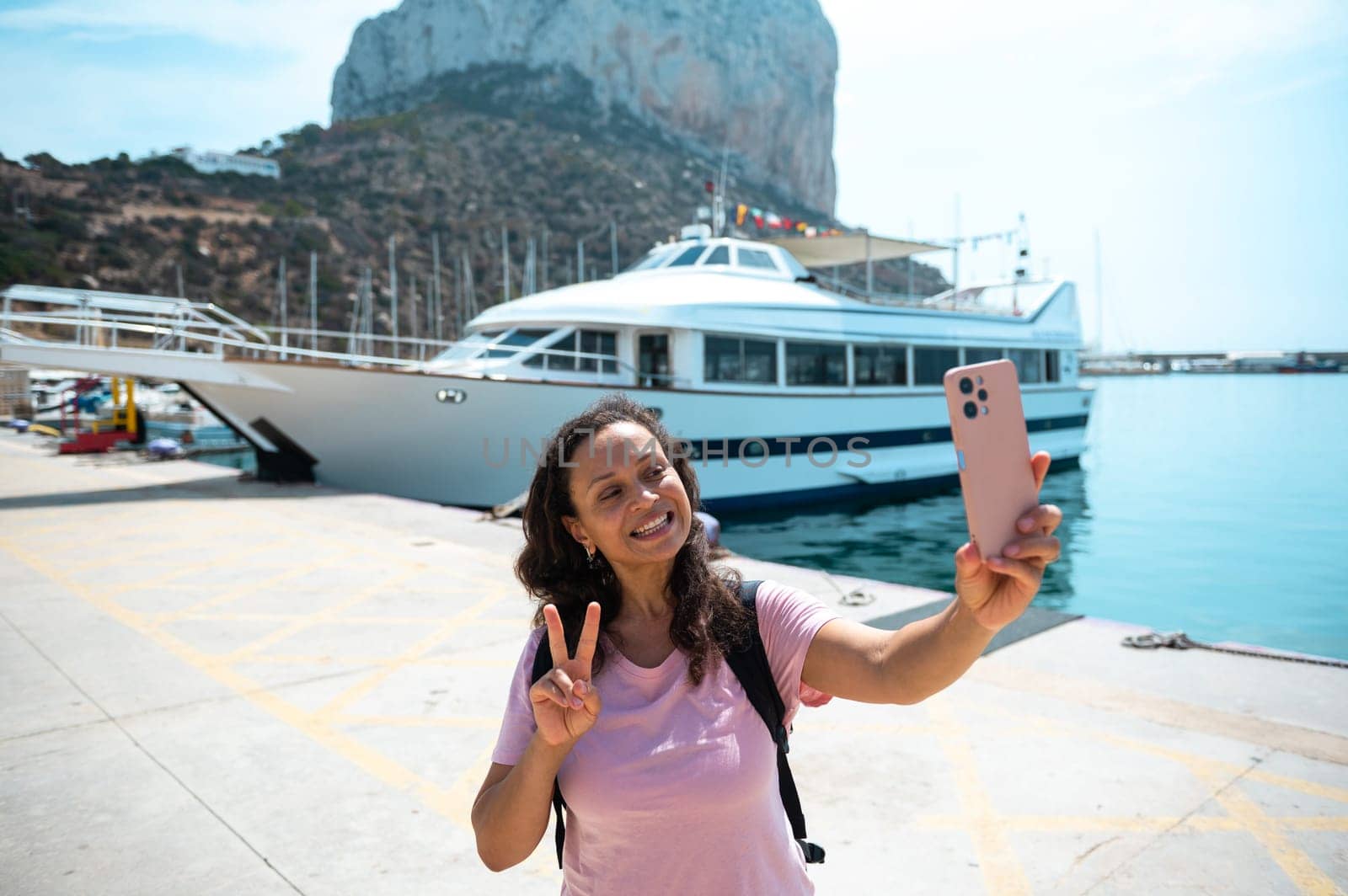 Smiling woman making a peace sign while taking a selfie near a yacht at a scenic harbor location.