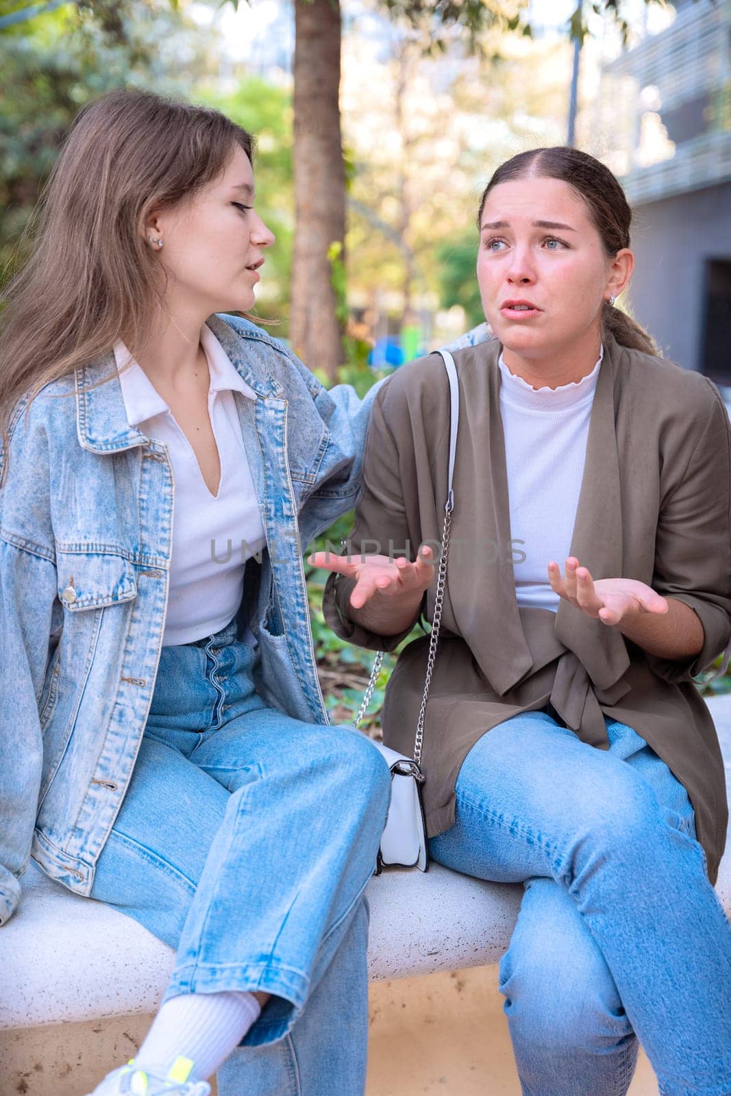 woman in a denim jacket comforting her sad friend who is crying. They are sitting in a park. The scene captures a moment of emotional support between friends. Vertical