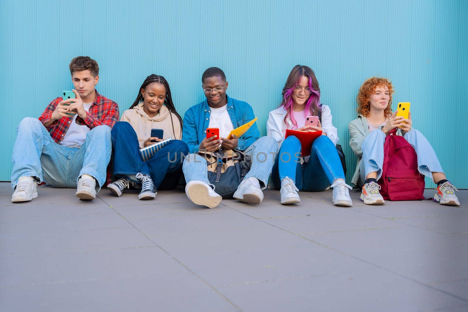 A diverse group of five smiling young students sit together on the floor, smiling and interacting on their smartphones against a blue background.