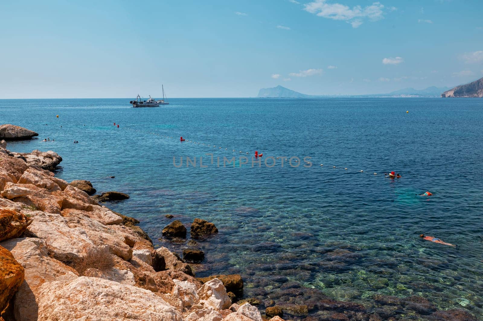 Beautiful coastal scene featuring clear blue waters, swimmers enjoying the sea, rocky shore, and a boat on the horizon under a clear sky.