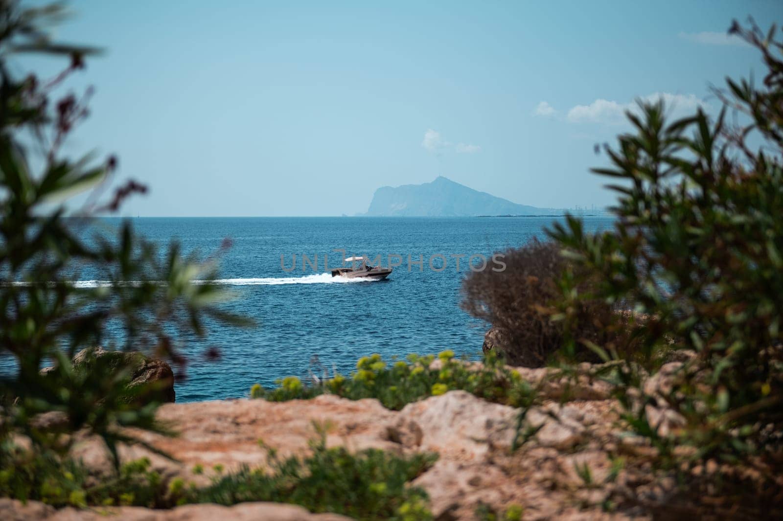 A speedboat glides across turquoise waters near a rocky coastline, framed by lush greenery and a distant island under a clear blue sky.