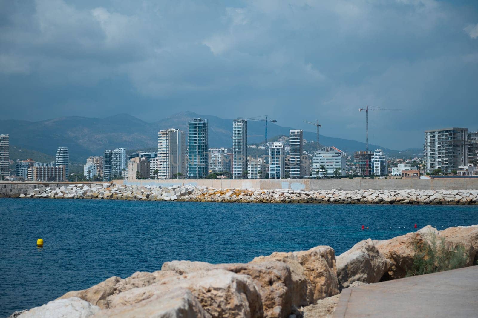A panoramic view of a coastal city featuring modern skyscrapers, calm blue waters, and mountains in the background, under a cloudy sky.