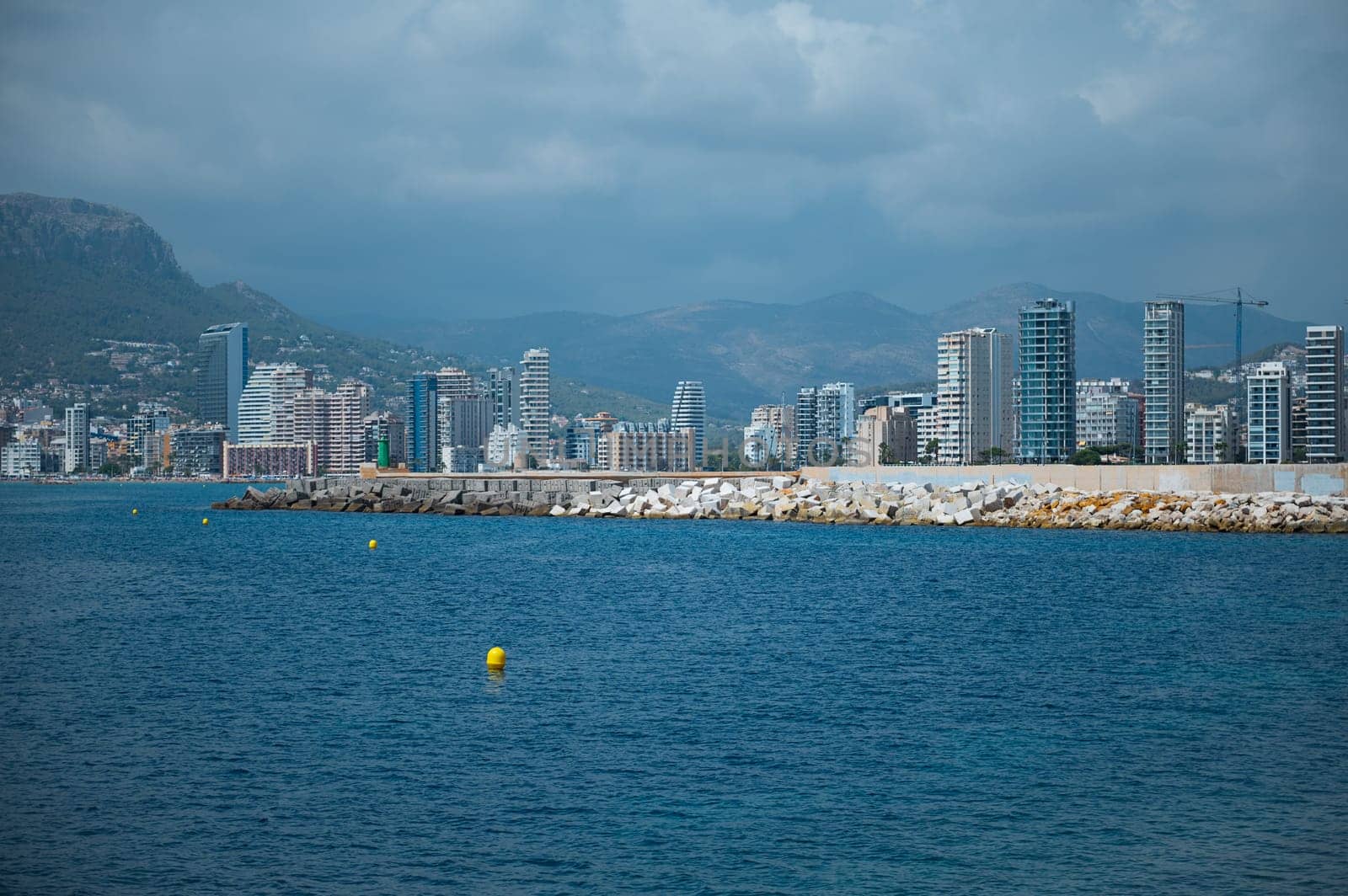 A vibrant coastal city skyline, against a backdrop of mountains under cloudy skies, reflecting urban development and nature beauty.