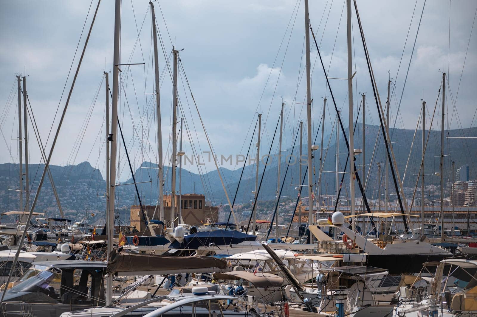A picturesque view of a busy marina filled with yachts against a backdrop of majestic mountains under a cloudy sky.