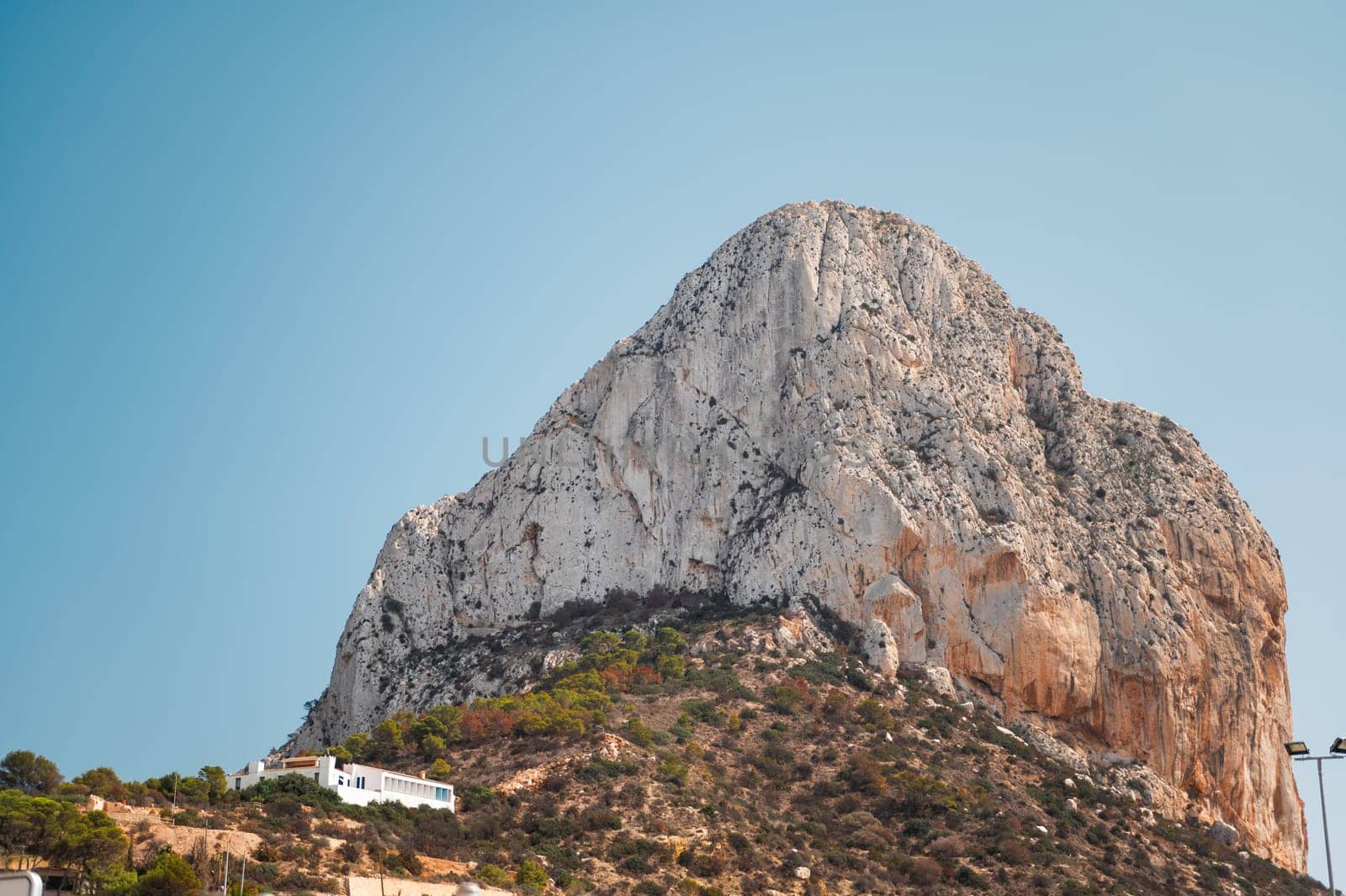 A towering rocky mountain stands majestically against a clear blue sky, with a small building nestled at its base, highlighting nature's grandeur.