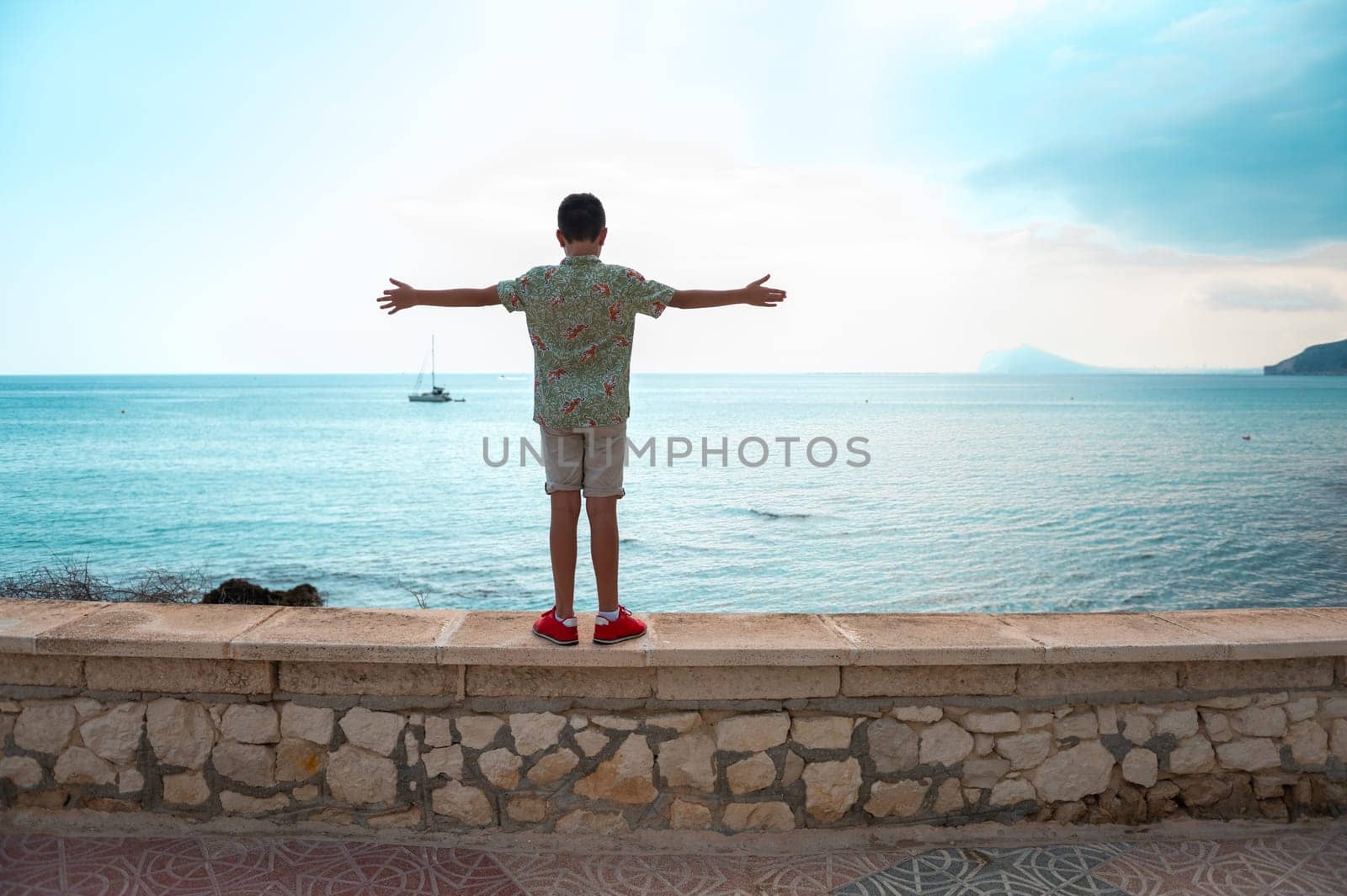 A young boy stands with open arms facing the sea, conveying a sense of freedom and adventure against a peaceful ocean backdrop.