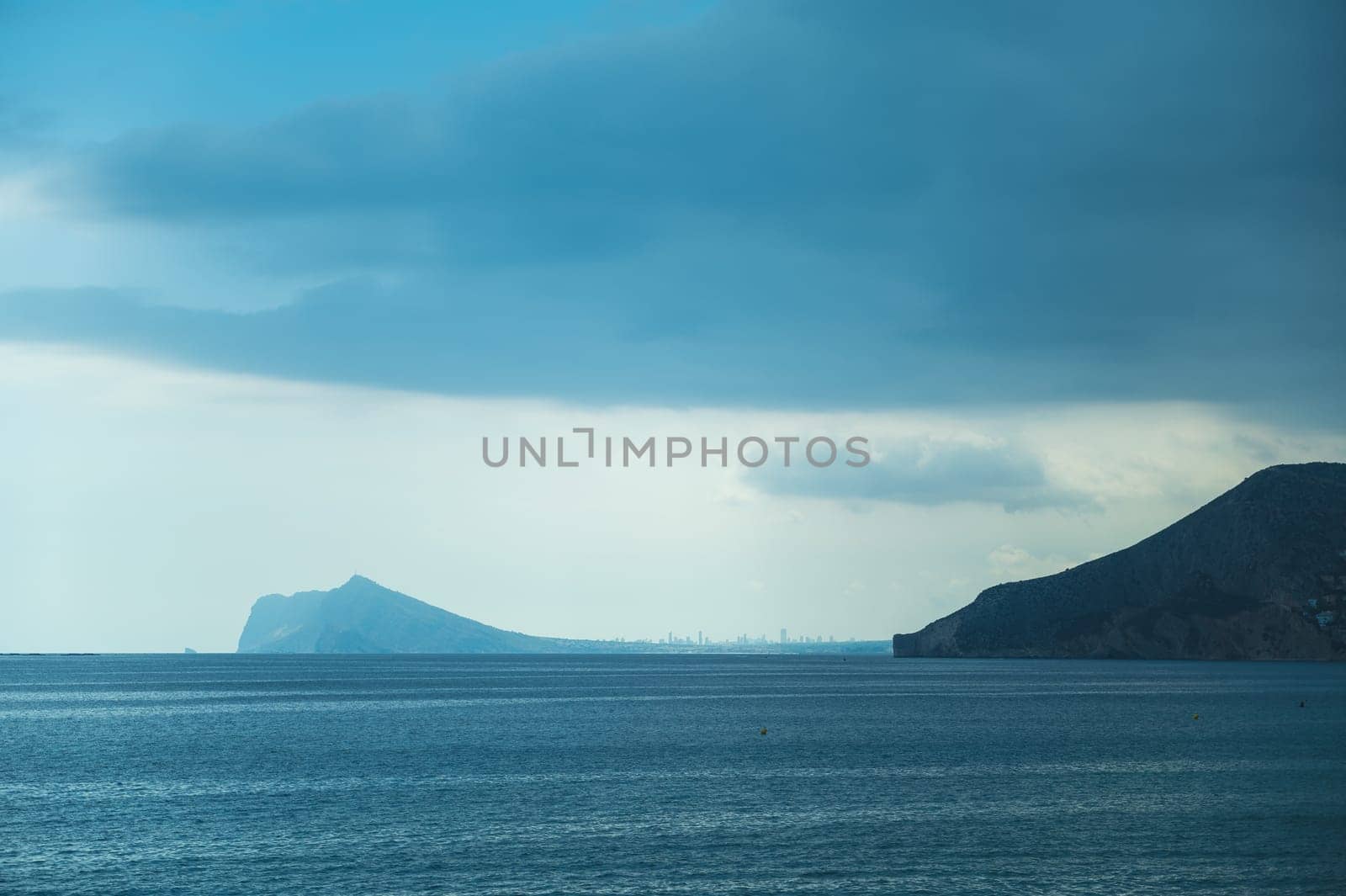 Peaceful seascape with the ocean under overcast sky, distant mountainous coastline, and city skyline on the horizon.