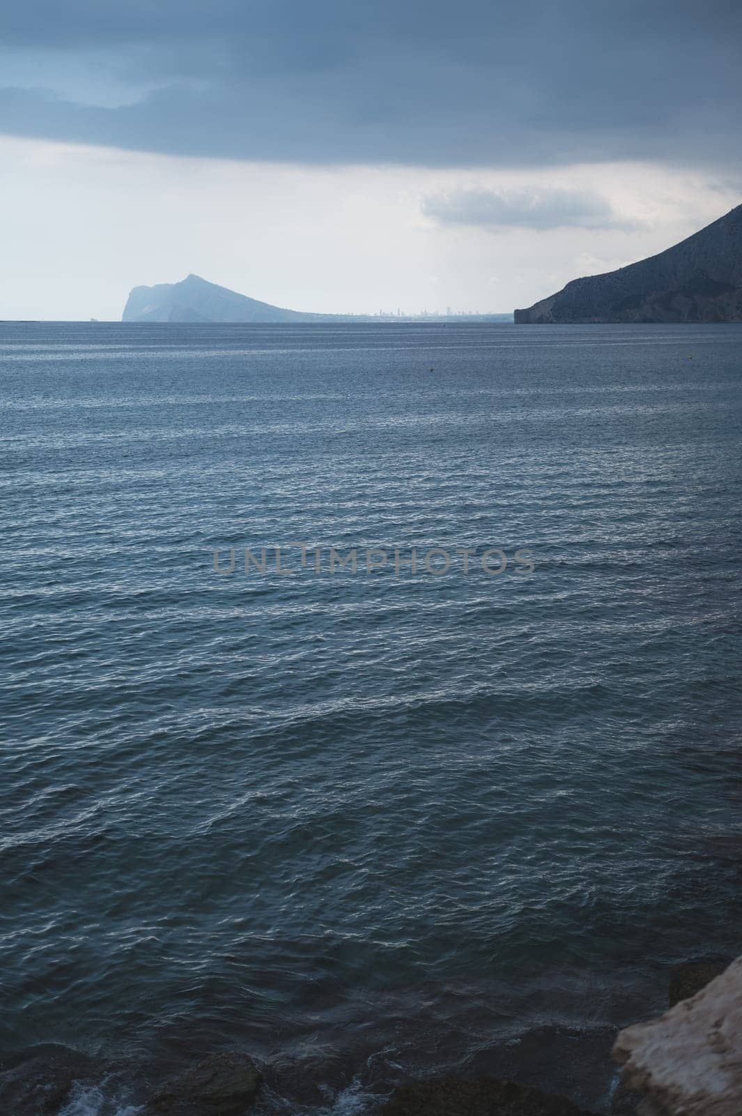 A tranquil seascape depicting calm blue waters under a cloudy sky, with distant mountains on the horizon, creating a serene and moody atmosphere.