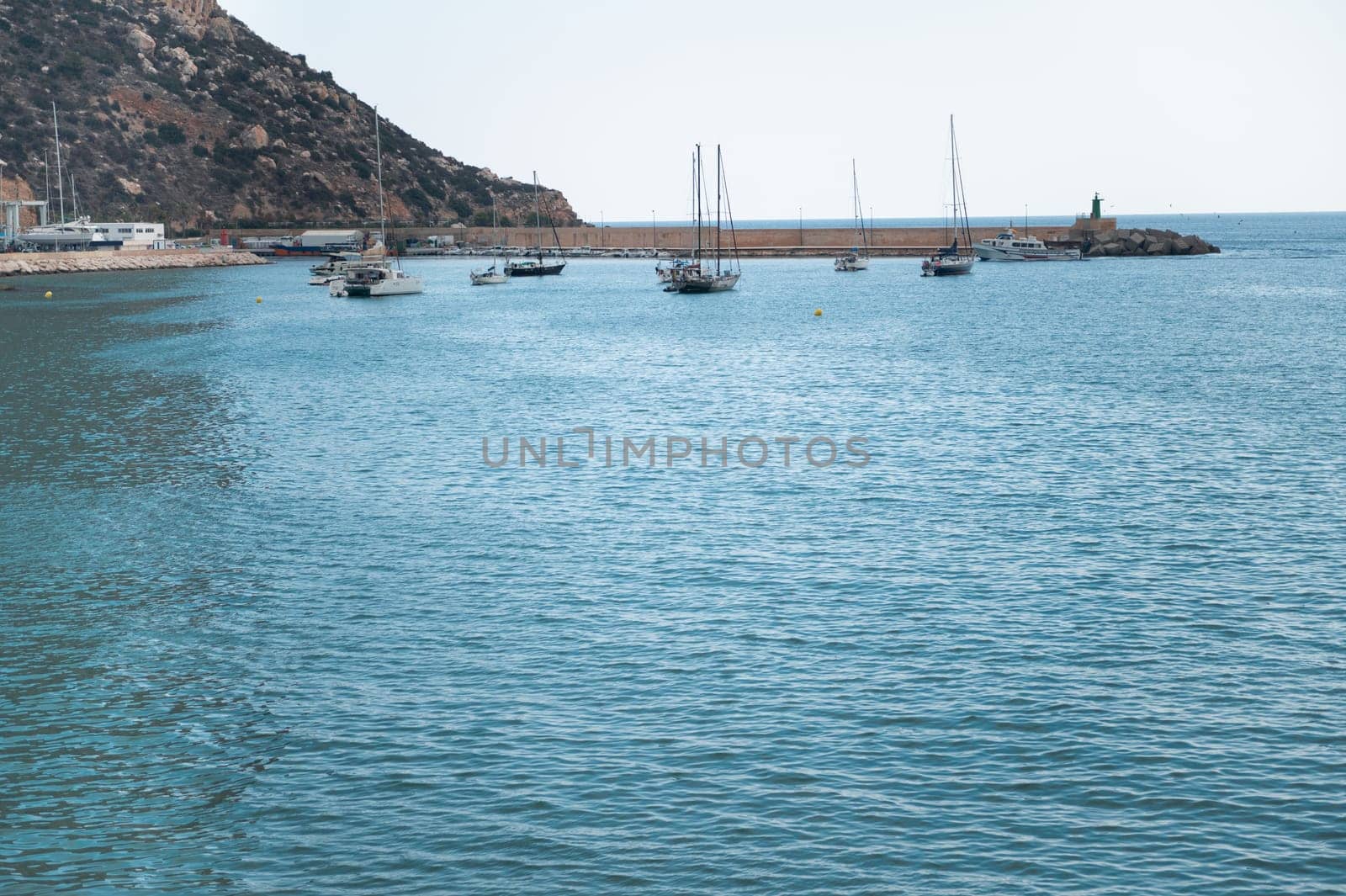 A peaceful coastal scene with several sailboats anchored in a calm marina, with a backdrop of a rocky mountain and open sea under a clear sky.