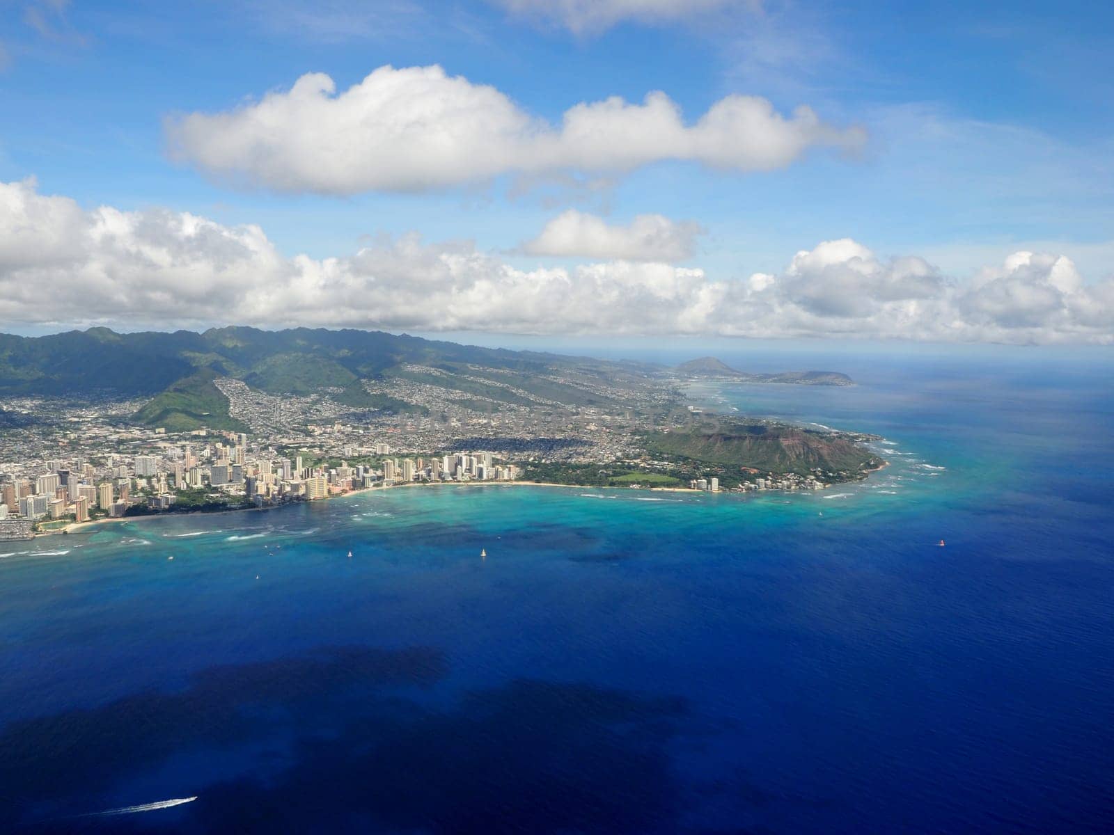 An expansive aerial perspective captures Honolulu’s stunning shoreline filled with turquoise waters, city buildings, and surrounding green mountains under a blue sky.