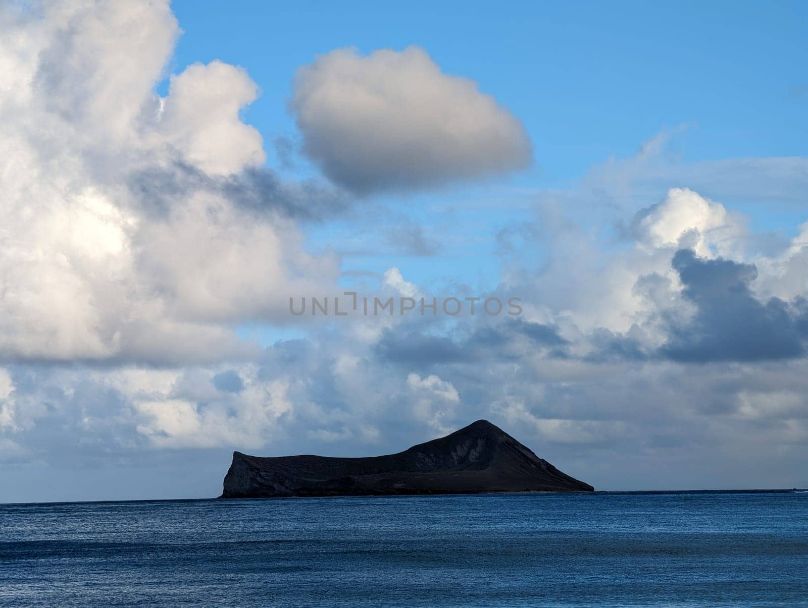 The image showcases a solitary island surrounded by calm waters, with a dramatic sky featuring soft, white clouds. A peaceful and scenic coastal vista invites tranquility.