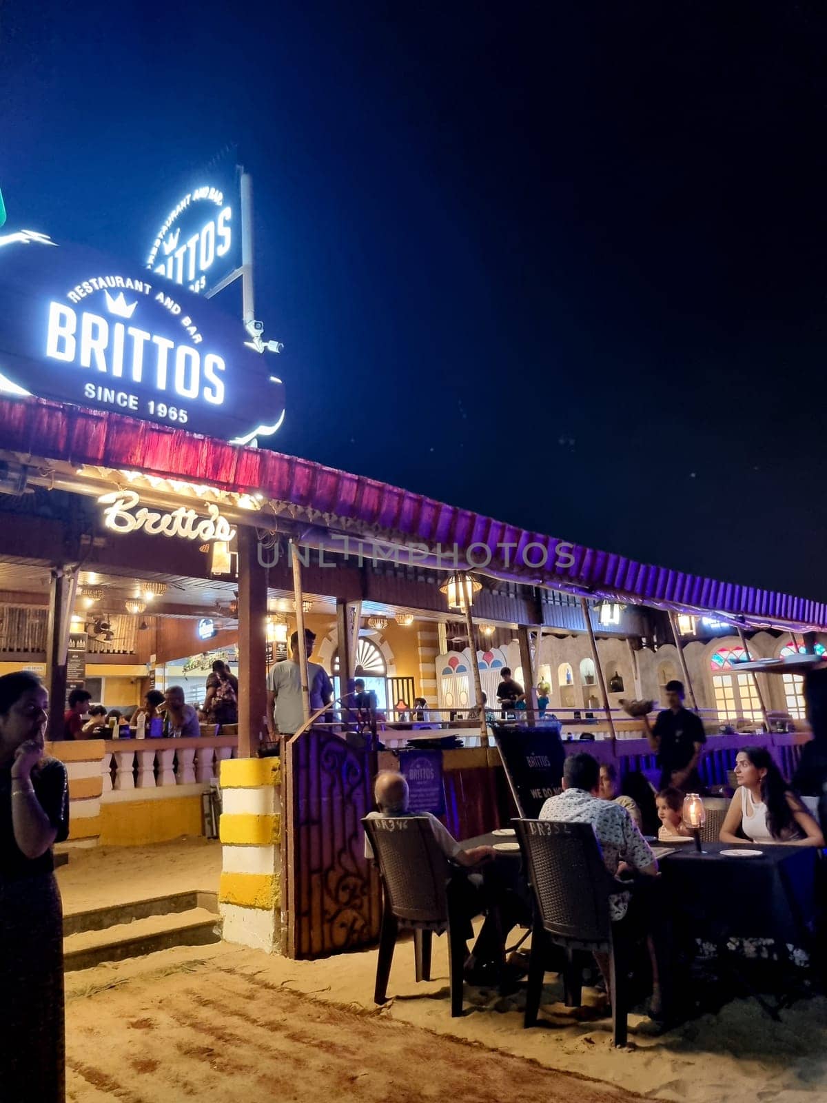 Goa, India - 16th Aug 2024: wide angle shot showing famous shack Brittos on baga beach with colorful lights and crowd of tourists enjoying dining on sand karaoke and more