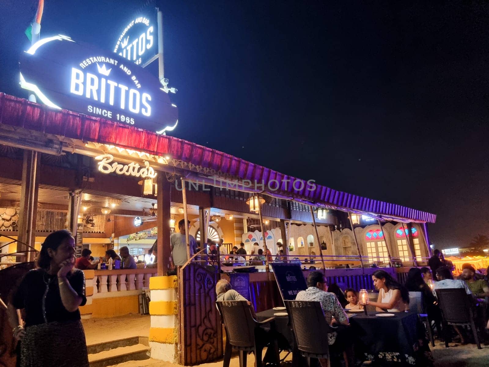 Goa, India - 16th Aug 2024: wide angle shot showing famous shack Brittos on baga beach with colorful lights and crowd of tourists enjoying dining on sand karaoke and more