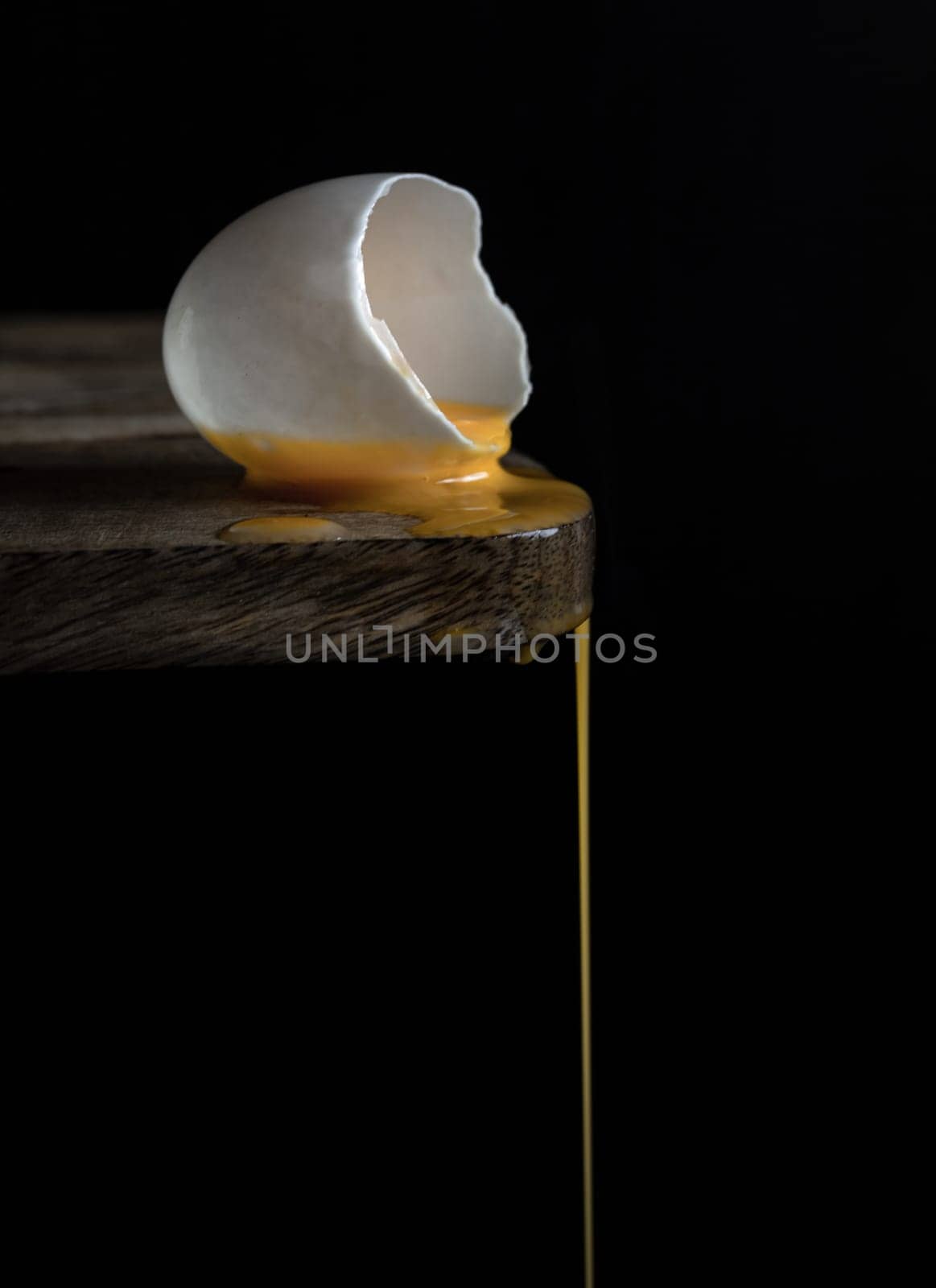 A cracked raw white duck egg shell and dripping yolk on the edge of a wooden cutting board and  black background. Conceptual image of a broken white eggshell with dripping egg yolk on dark background, use it as your Wallpaper, Poster and Space for text, Selective focus.