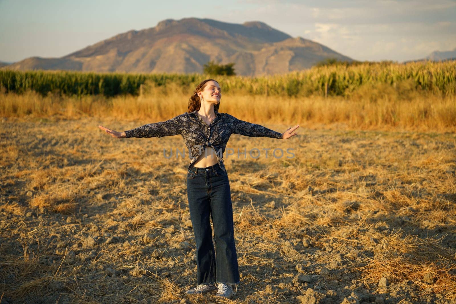 A young woman stands with her arms wide open in a beautiful golden field, completely surrounded by majestic mountains, radiating a deep sense of freedom, happiness, and pure joy in this moment