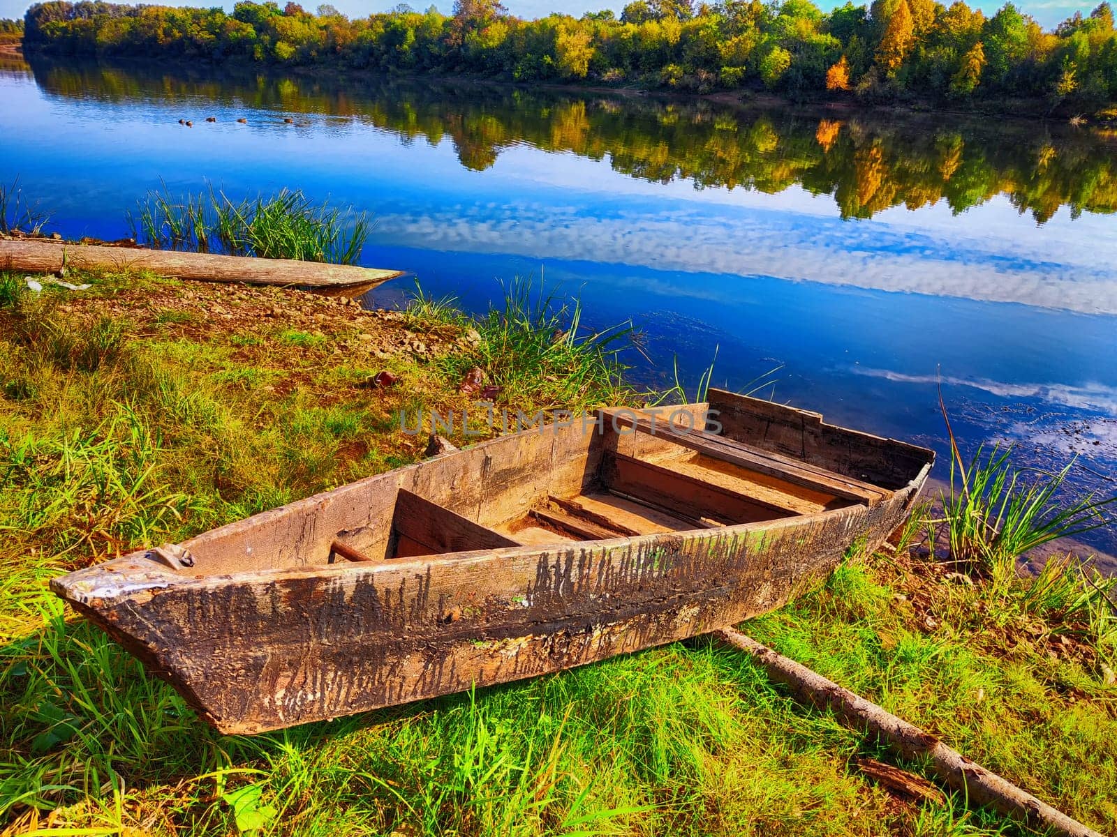 A weathered boat sits on the riverbank lush with green grass and vibrant fall colors reflecting on the water.