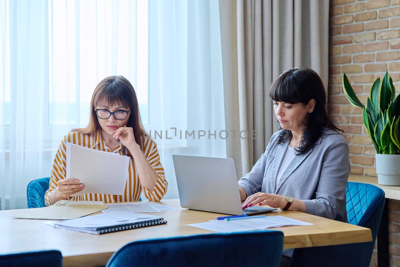 Two mature business women working in office, with papers, laptop by VH-studio