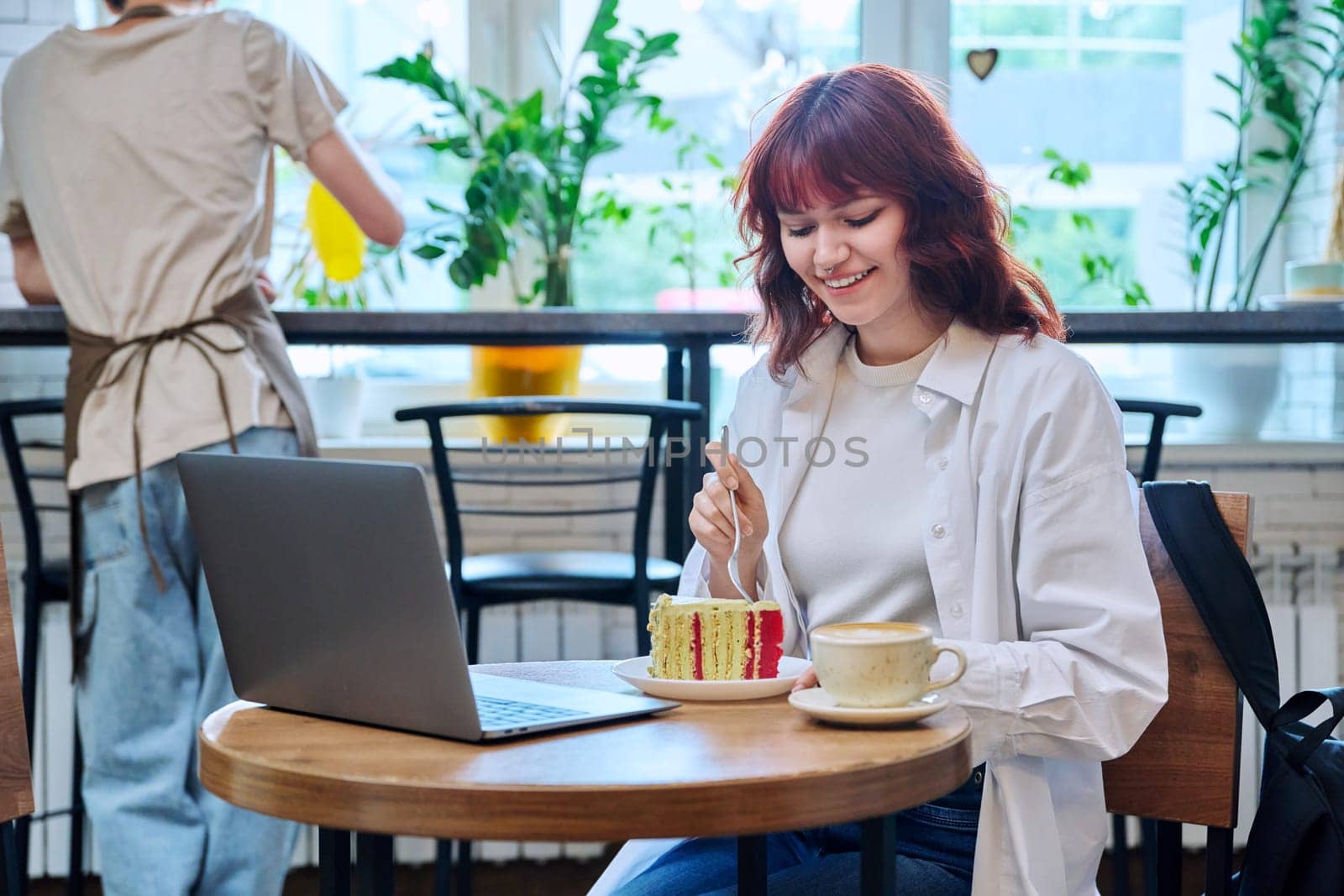 Female college student with laptop in cafeteria at table with cup of coffee and piece of cake. Internet online technology for leisure communication blogging learning chat, youth lifestyle concept