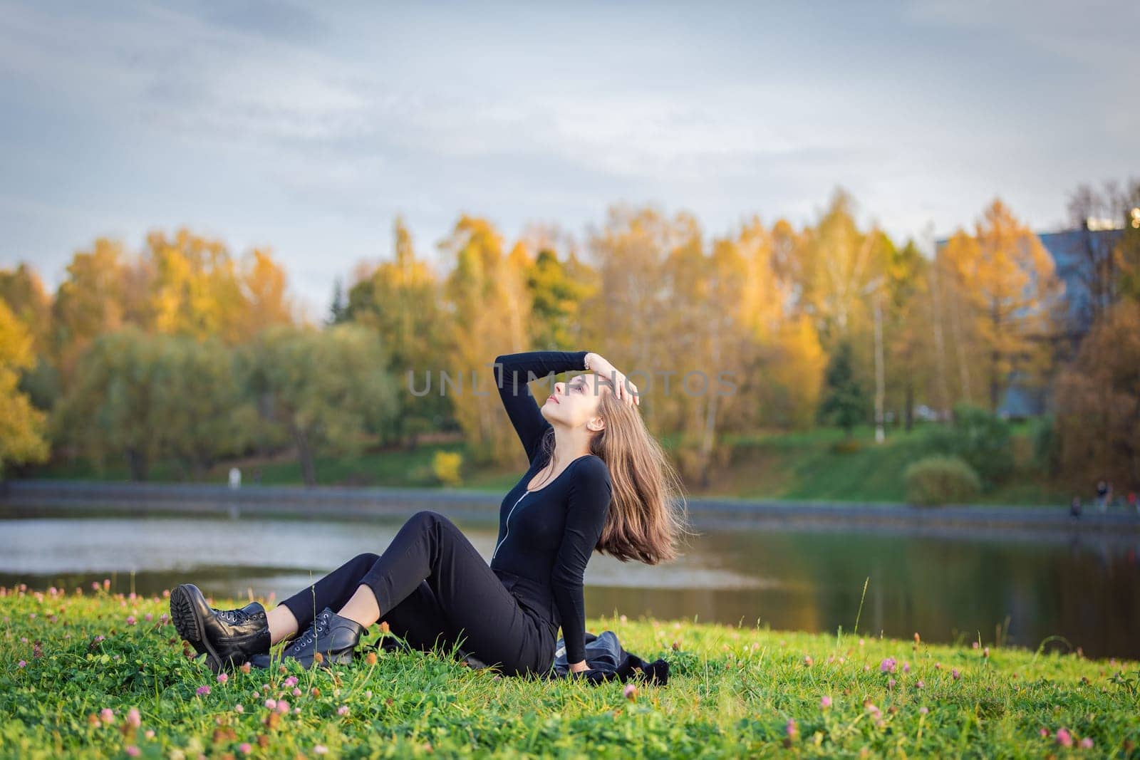 A beautiful girl poses while sitting on the grass by a pond in an autumn park