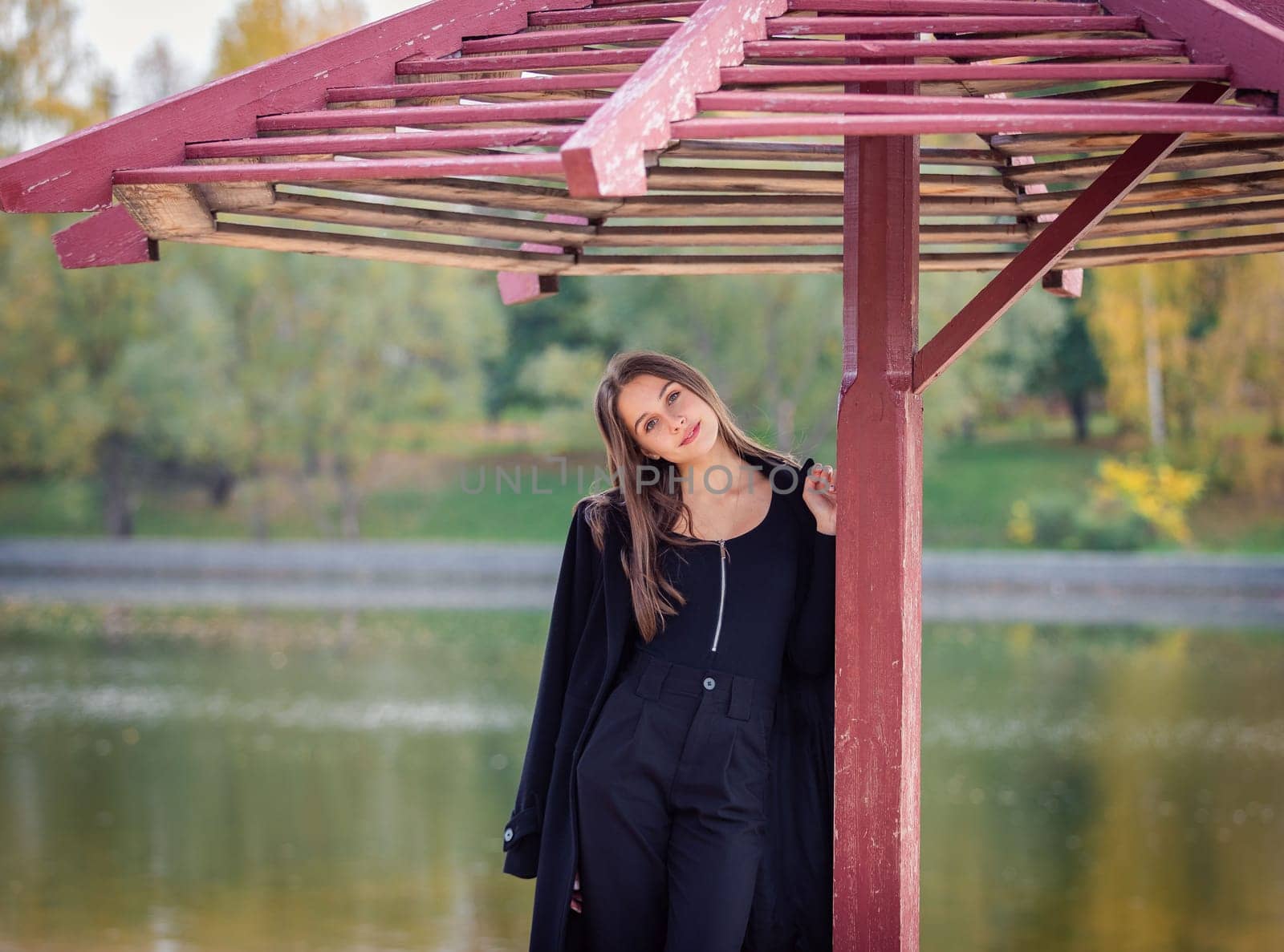 A beautiful girl poses while standing by a pond under an umbrella in an autumn park