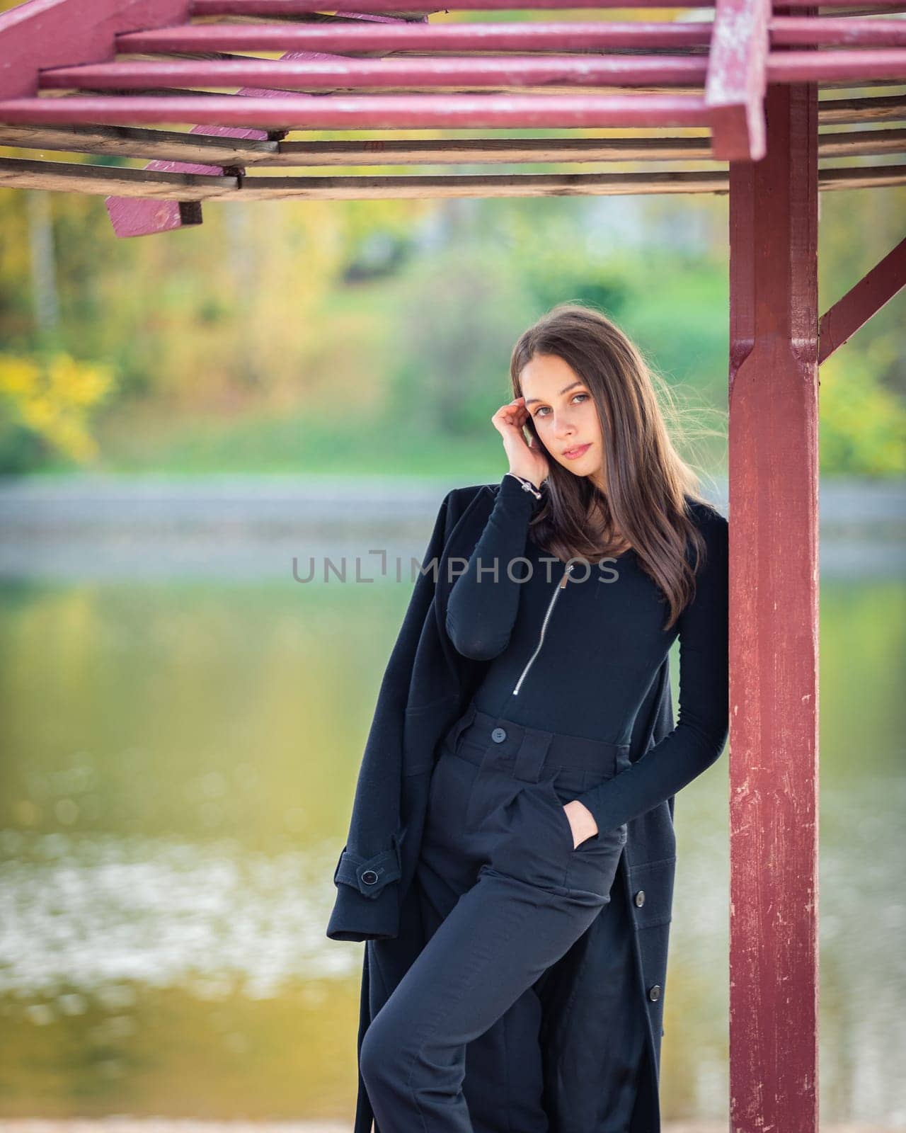 A beautiful girl poses while standing by a pond under an umbrella in an autumn park