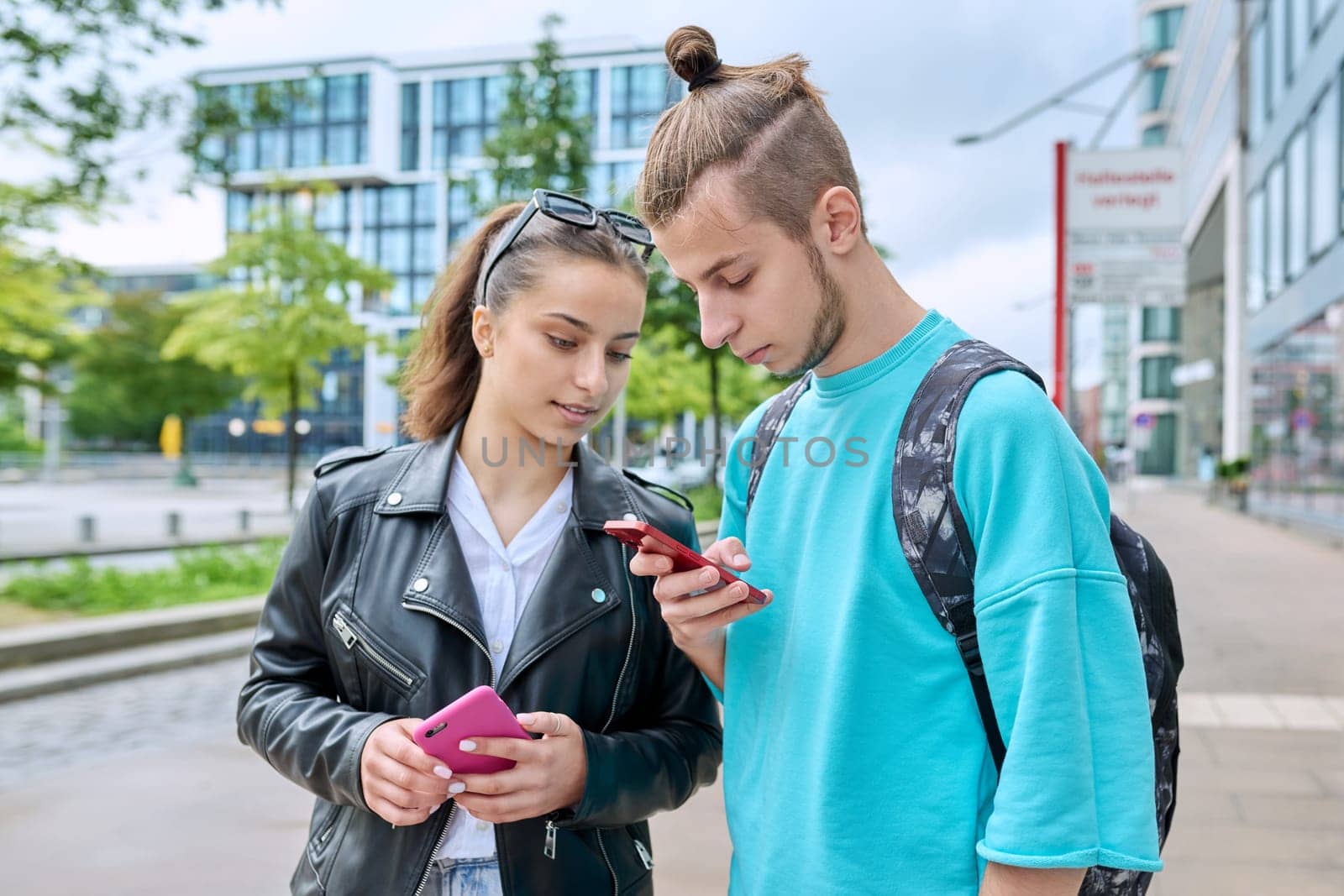Teen friends guy and girl standing together holding smartphones looking at screen using mobile phones outdoor on city street. Internet digital technology applications for leisure study communication